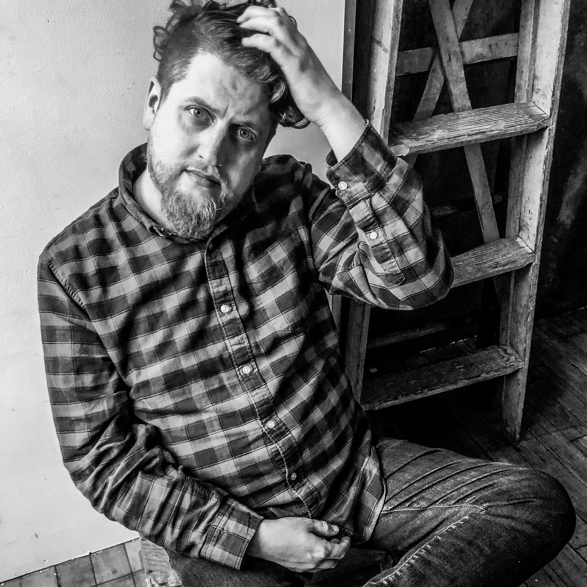 Black and white photo of a man with a beard and mustache, wearing a plaid shirt, sitting on the floor with a distressed expression, holding his head with one hand, next to a wooden chair or stool.