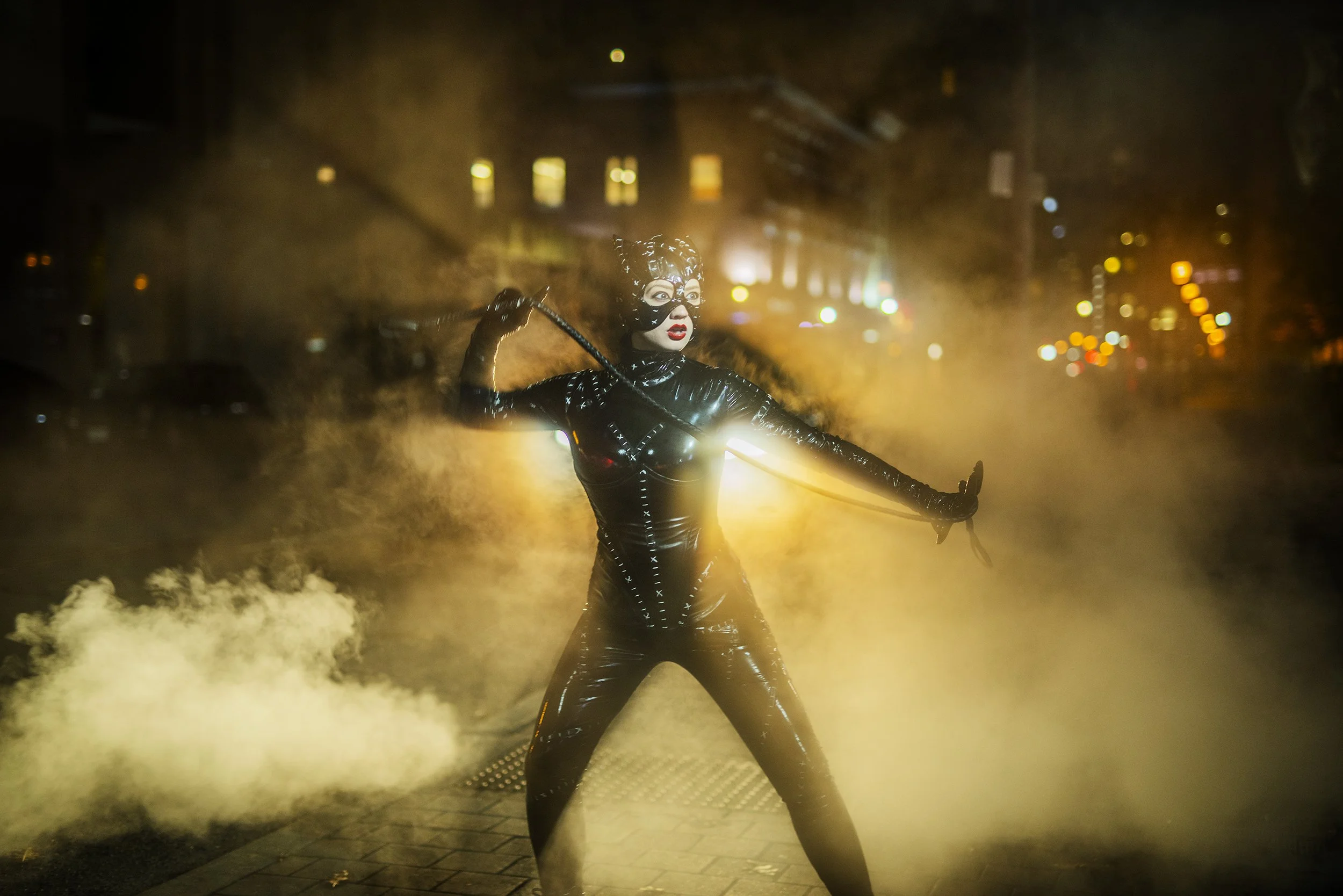 A person dressed in a shiny black outfit with glasses and red lipstick, holding a stick, standing amidst yellowish smoke or mist on a city street at night with blurred city lights in the background.
