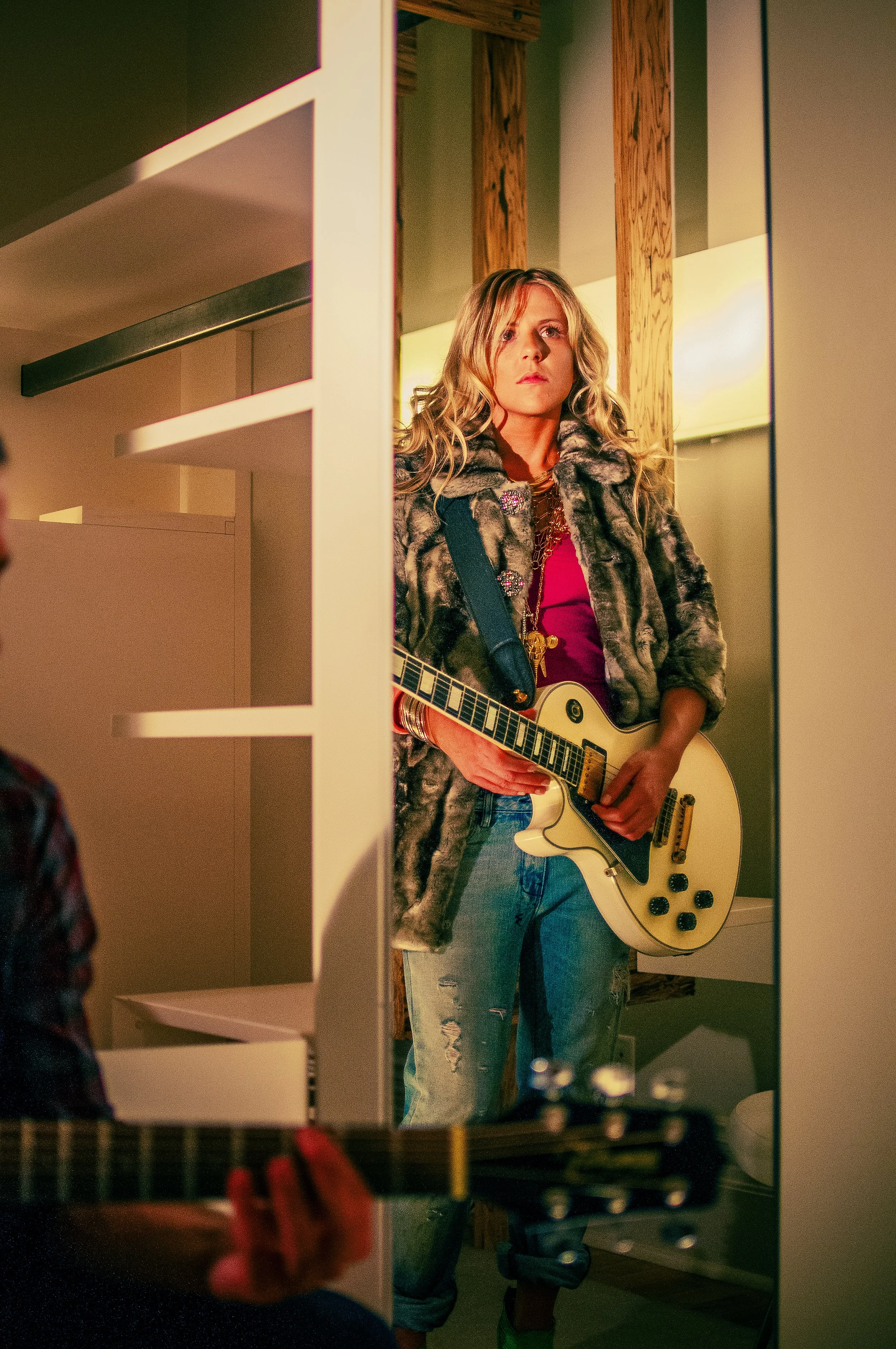 A young woman with wavy blonde hair is standing in a room, looking at her reflection in a mirror while holding a cream-colored electric guitar. She is wearing a patterned fur coat, a red top, and jeans.