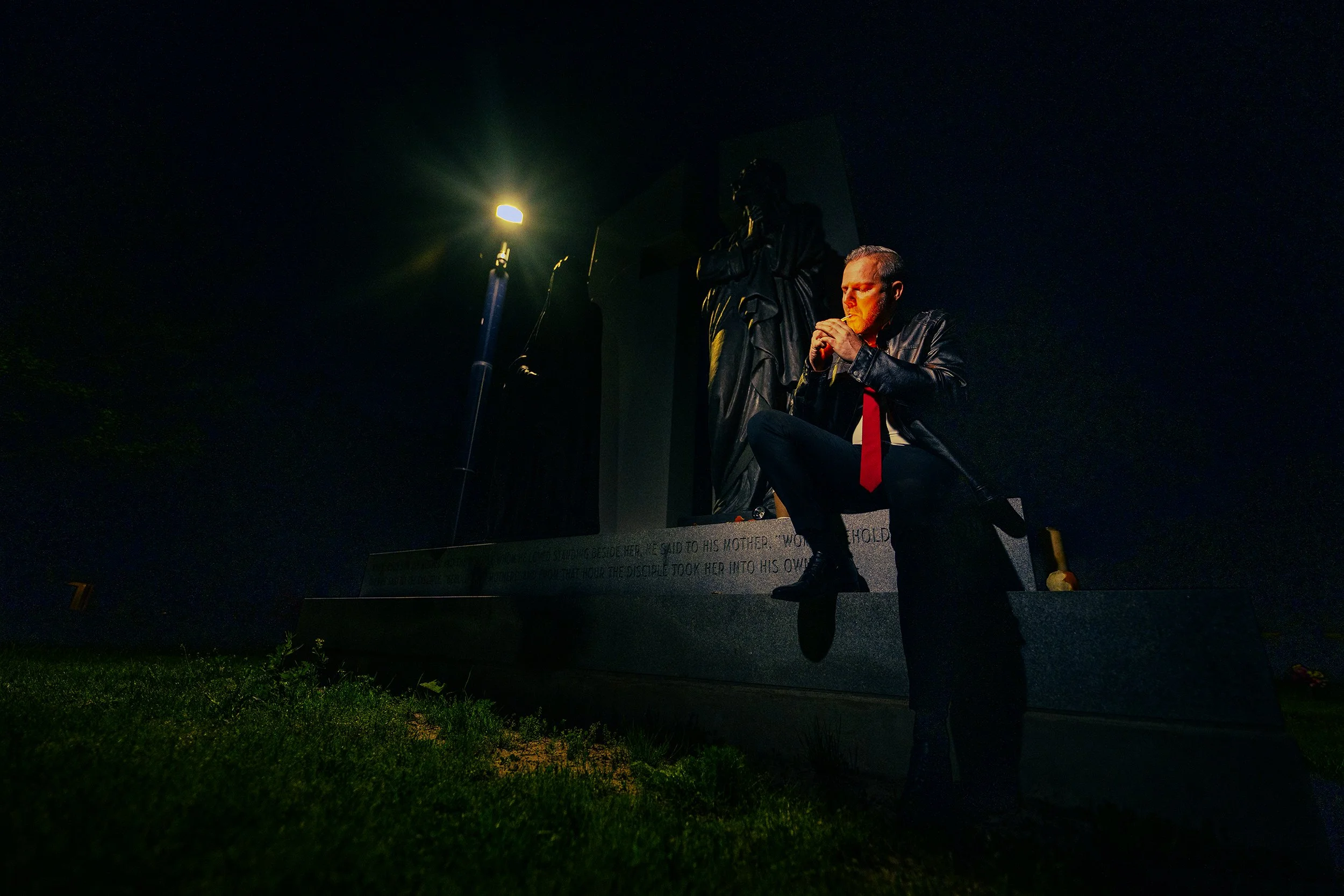 Nighttime scene of a man sitting on steps with a candle and a religious statue in the background, lit by a streetlamp.