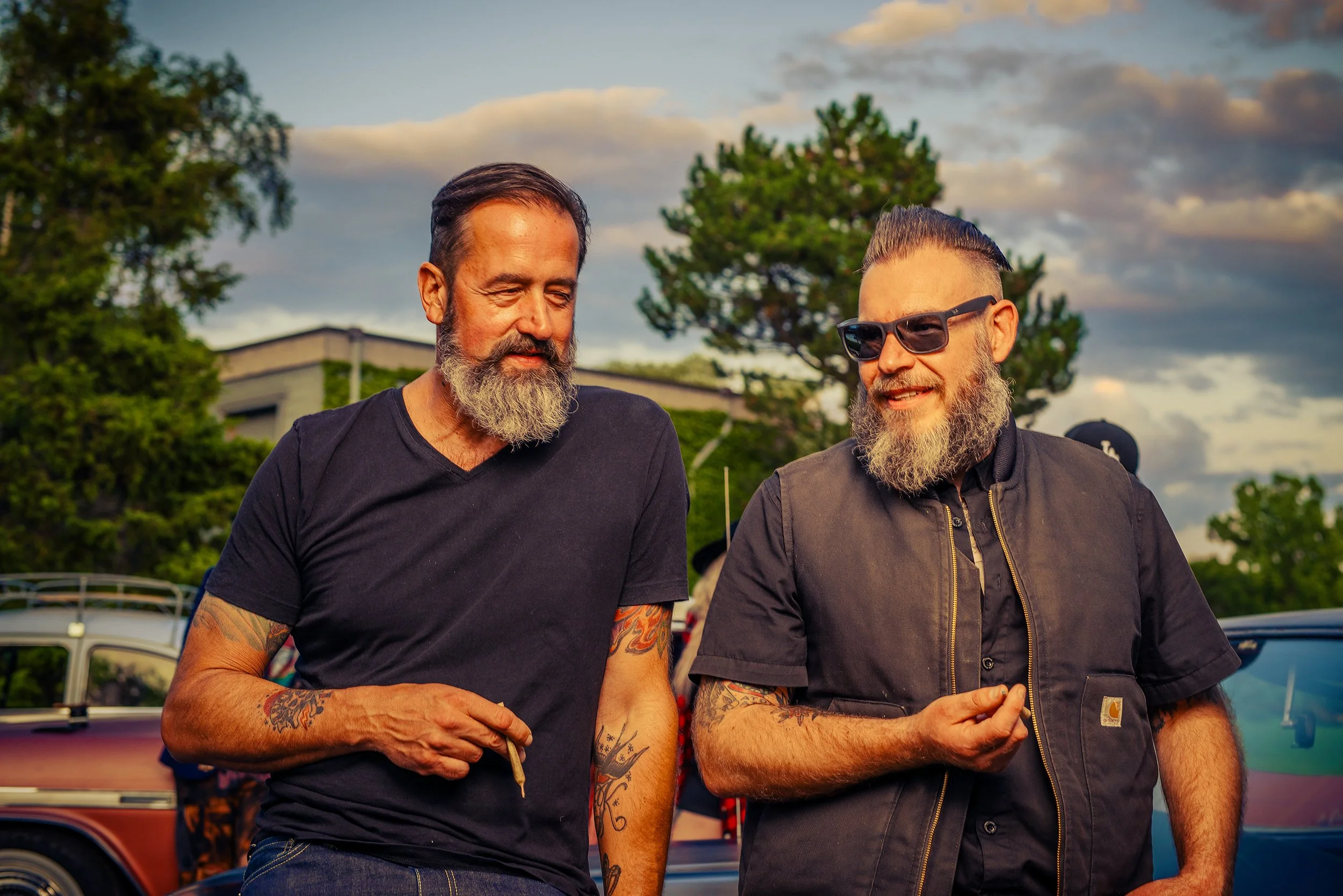 Two men with beards and tattoos outdoors at sunset, engaging in conversation, with trees and boats in the background.