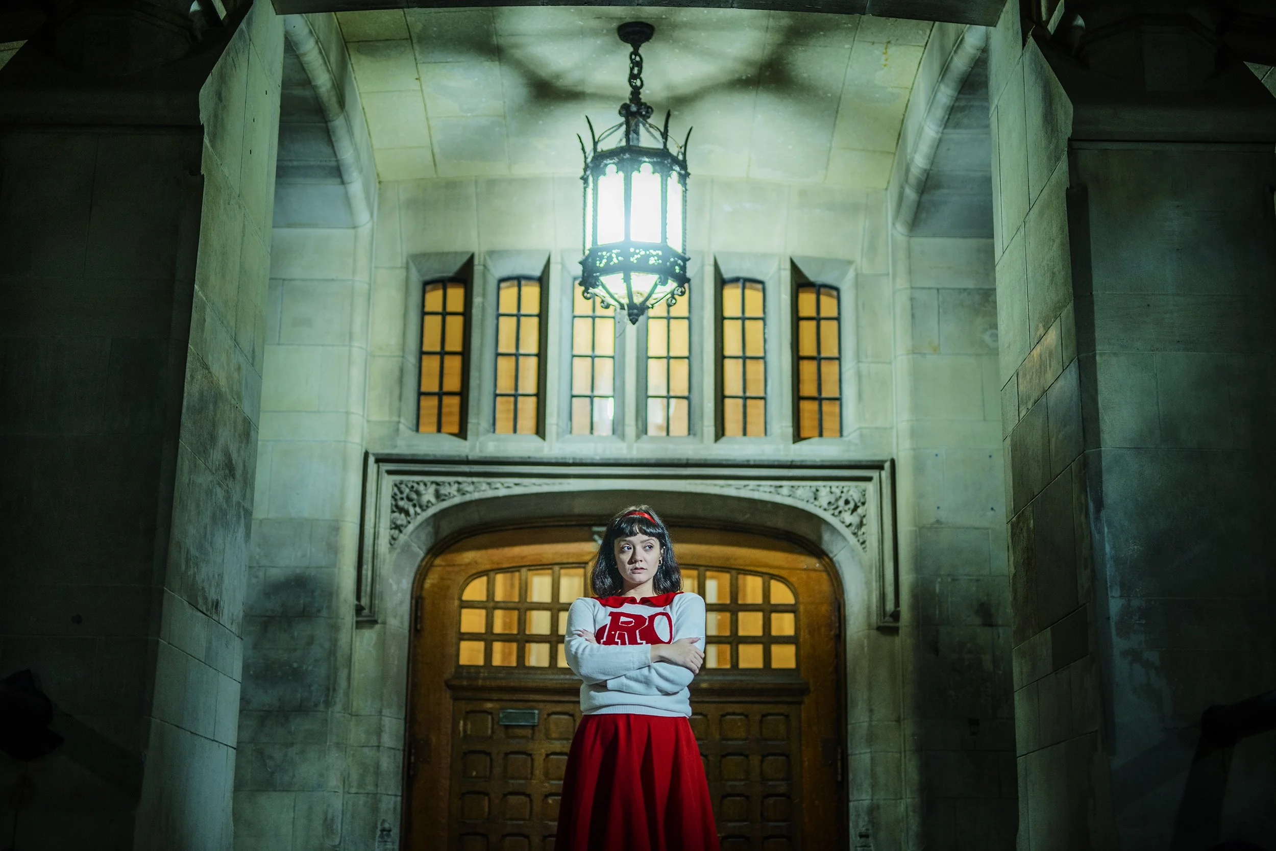 A woman with dark hair and bangs, wearing a white sweater with a red letter R on it and a red skirt, standing with her arms crossed in front of a large wooden doorway inside a stone building with a high ceiling and a chandelier.