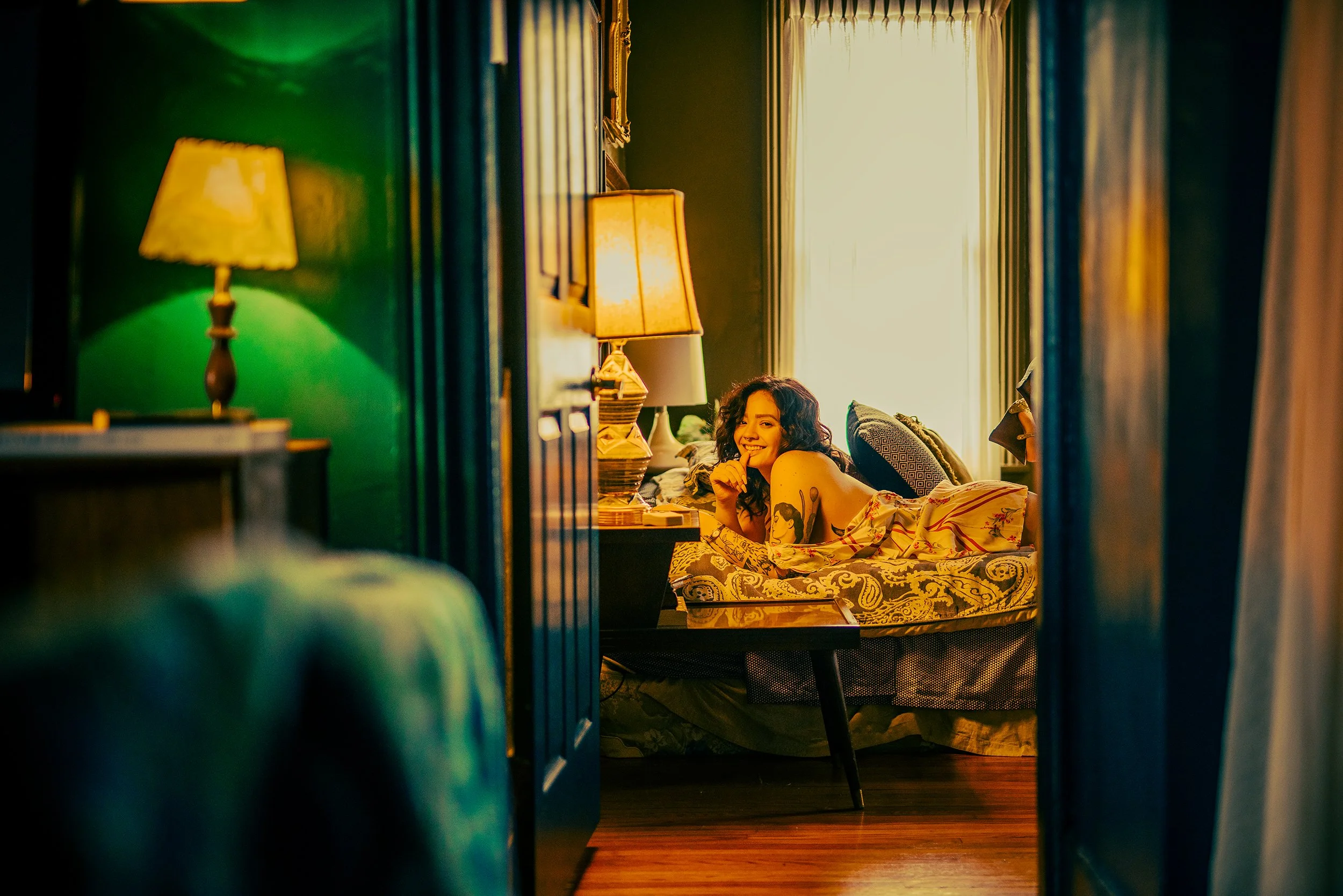 A woman with curly dark hair and tattoos lying on a bed, smiling, in a warmly lit bedroom with green walls and large windows, viewed through a doorway.