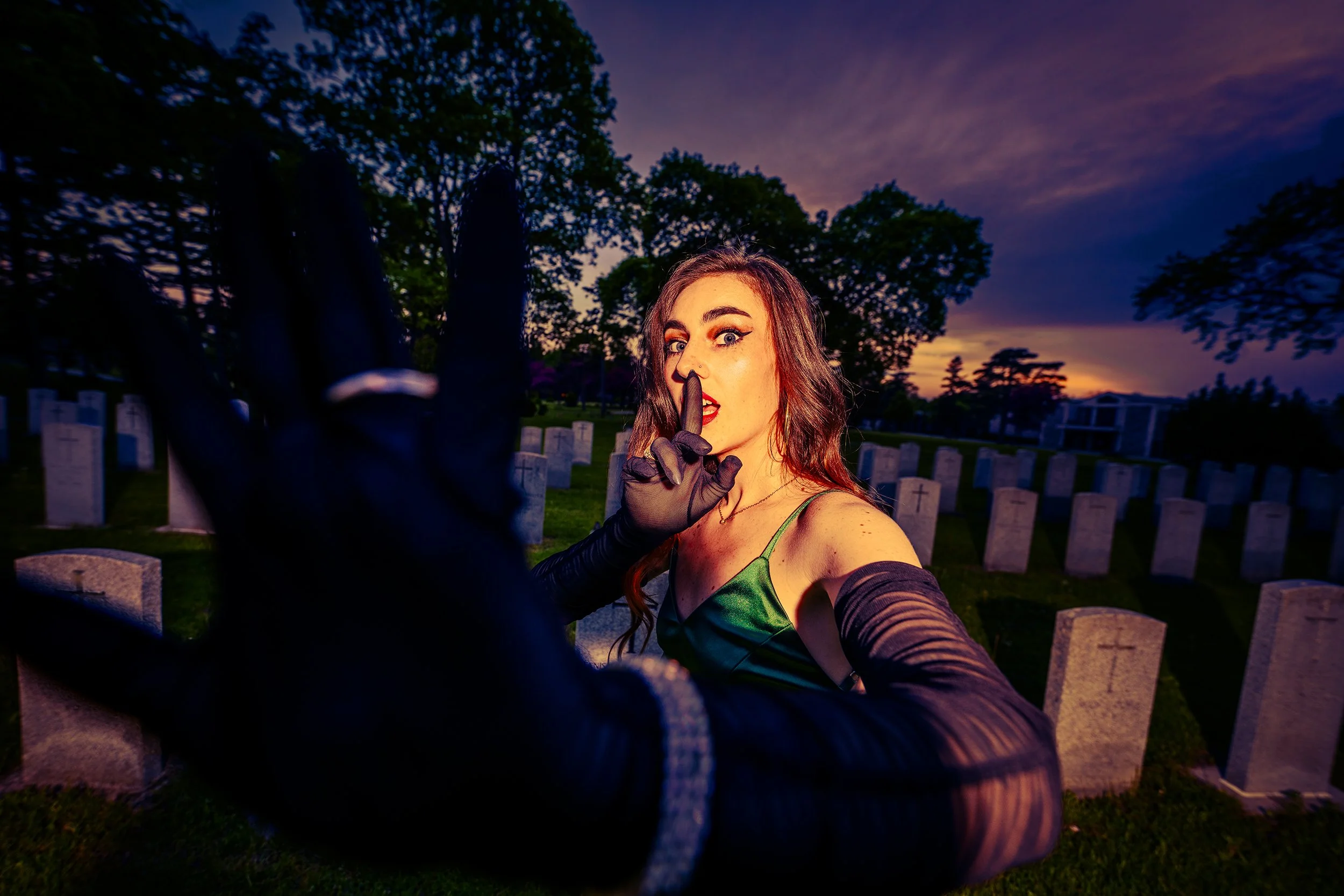 A woman with long, wavy hair and bold makeup, dressed in green and black, is taking a selfie in a graveyard at sunset. She has her finger to her lips in a shushing gesture and is wearing black gloves. The background features headstones and trees unde