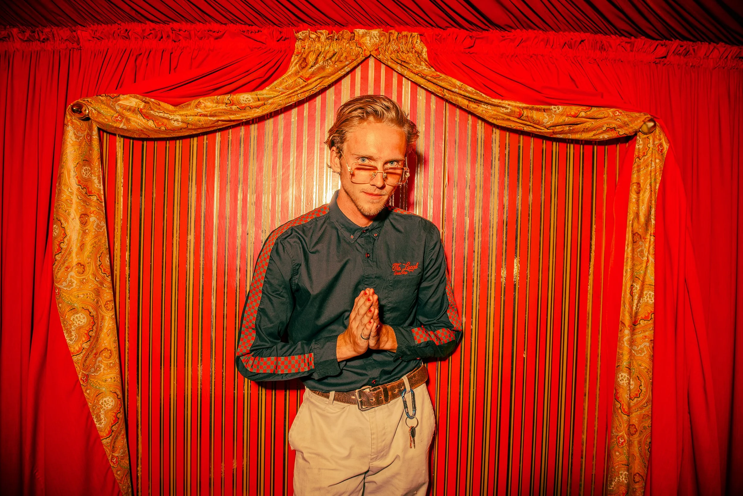A man with glasses, wearing a black shirt with red checkered accents, standing with hands in a prayer position in front of a red and gold curtain backdrop.