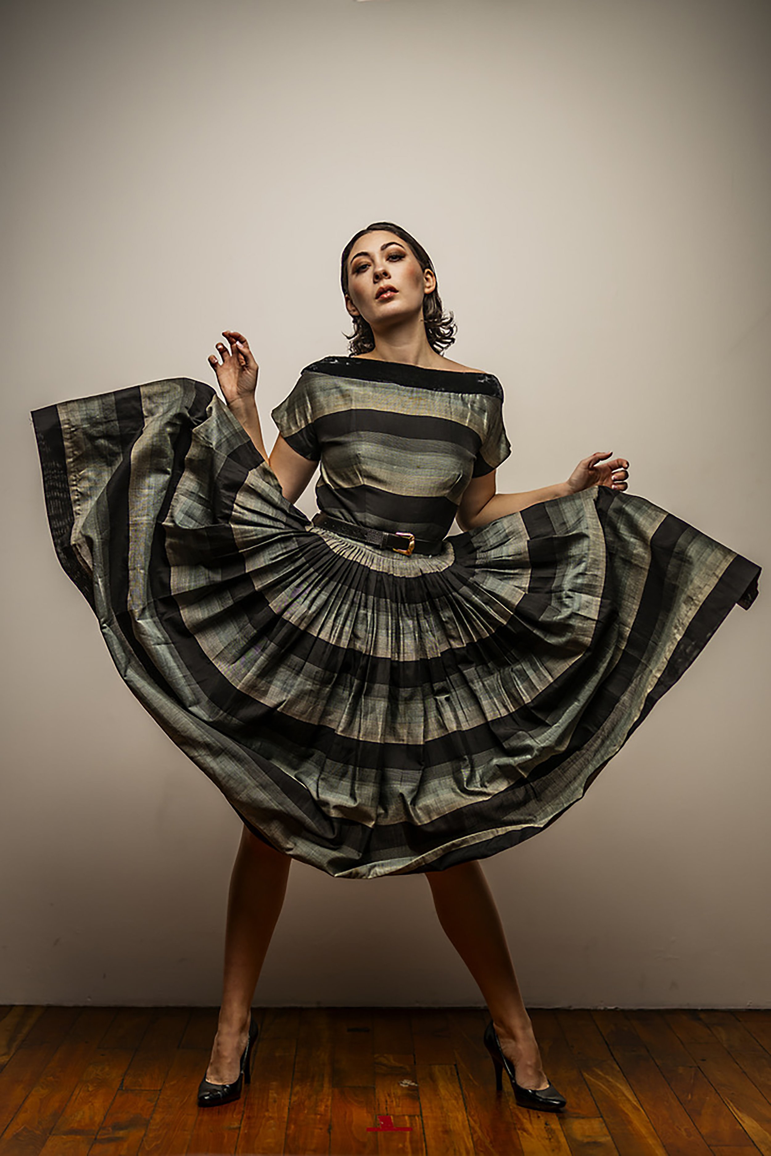 Woman in a plaid dress with a full skirt, standing with legs apart and arms raised, against a neutral background and wooden floor.