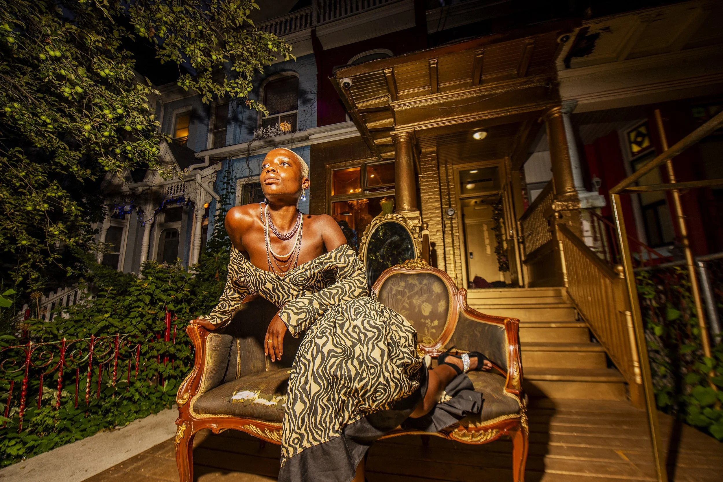 A woman sitting on an ornate vintage sofa outside at night, wearing a zebra print dress and jewelry, with a house and garden in the background.