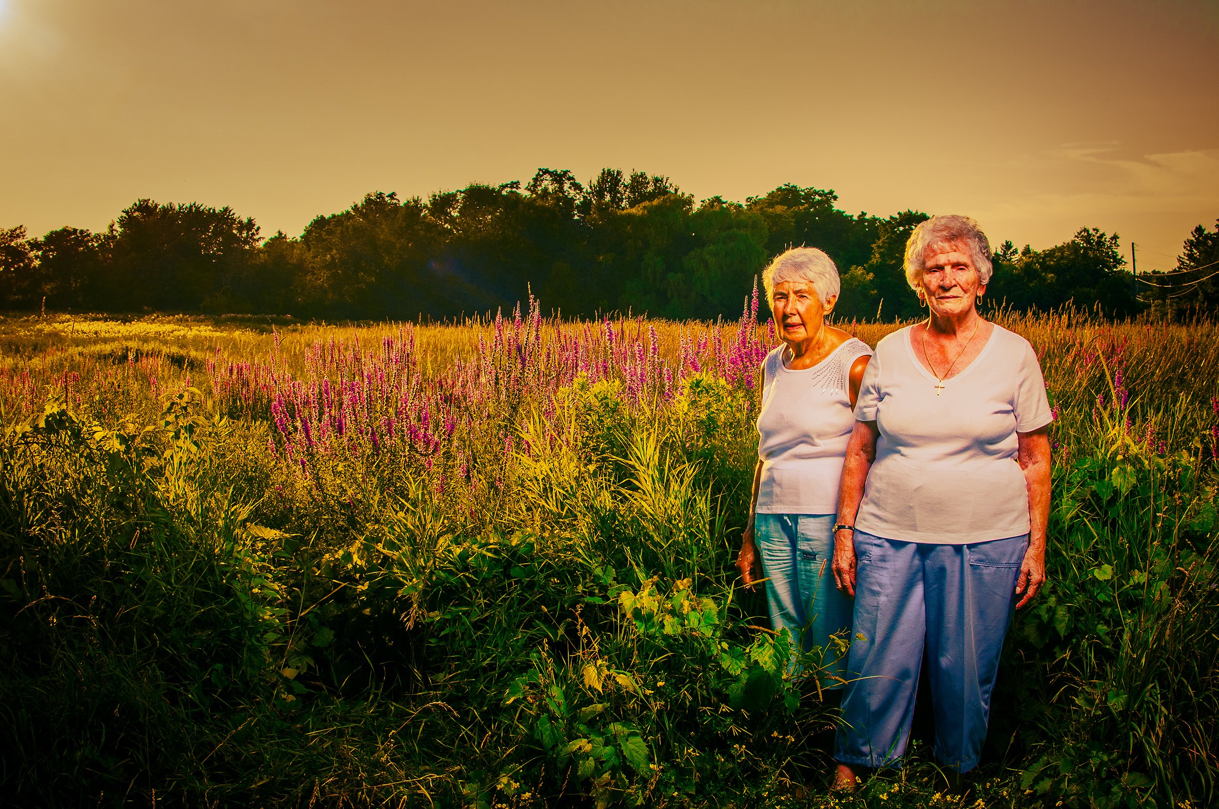 Two elderly women standing in a field of tall grass and purple flowers with trees and a sunset sky in the background.