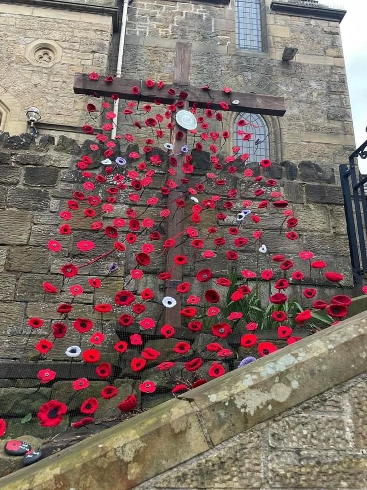 A large display of red poppies attached to a cross on a stone wall, located outside a stone building.