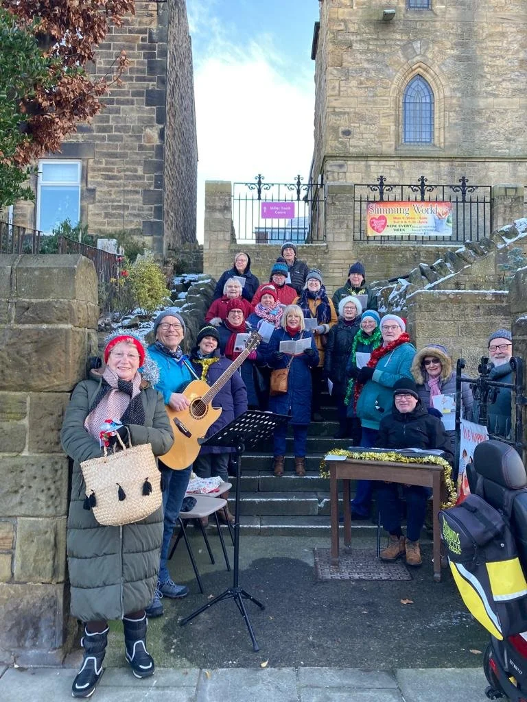 Group of people gathered on outdoor stone steps, some holding musical sheets, with a person playing guitar, in front of old stone buildings and banners celebrating Christmas.