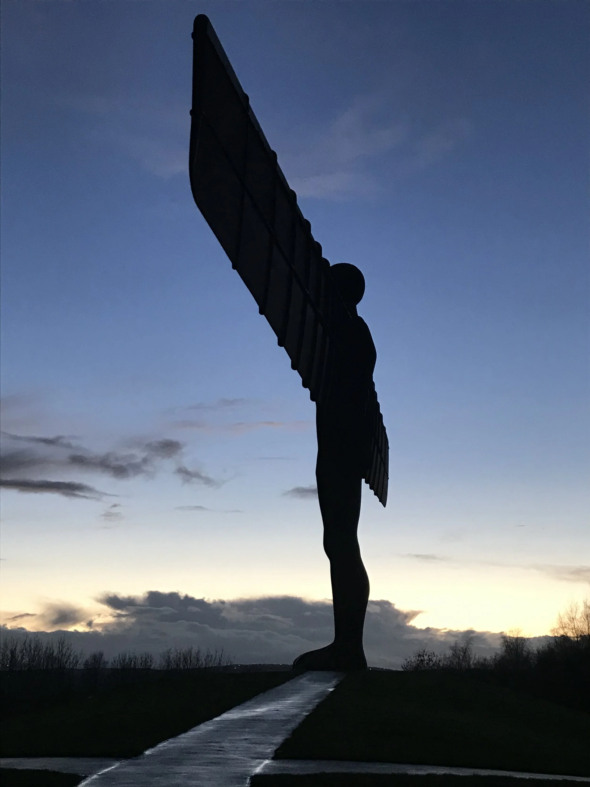 Angel of the North. Silhouette of a large statue of a figure with wings, standing on a hill at sunset or dusk, with a pathway leading up to it.
