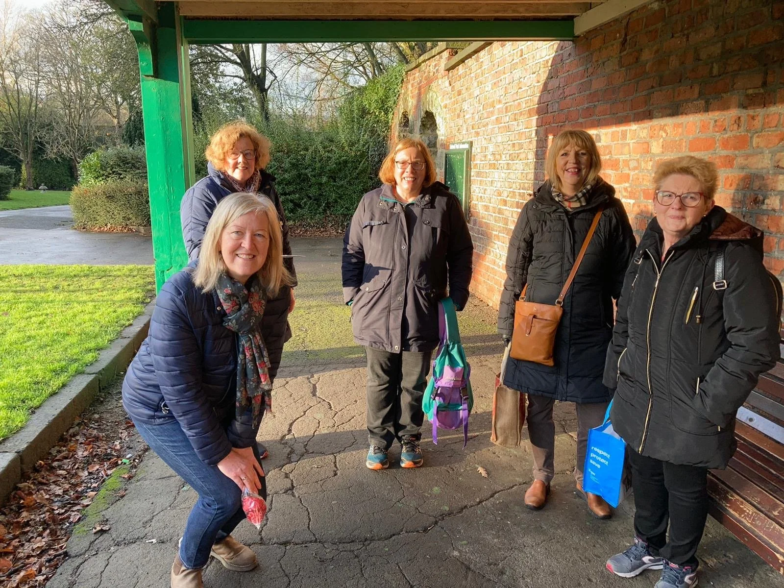 Six women standing outdoors on a cracked pavement near a brick wall, smiling at the camera during daytime. They are dressed in jackets, some carrying bags or backpacks, with trees and greenery in the background.