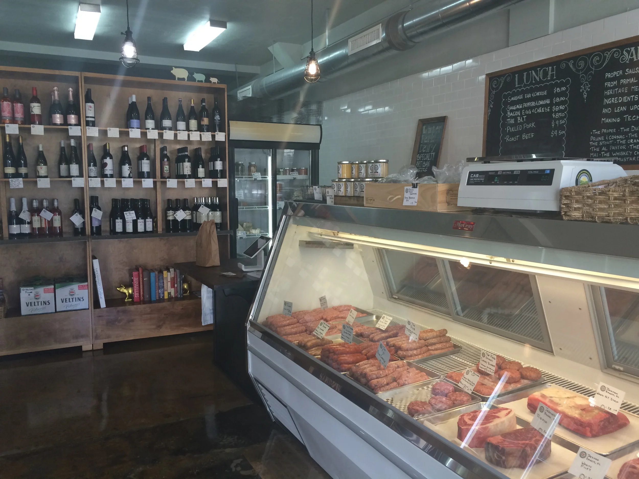 O refrigerador de carnes e embutidos, com a adega de vinhos inusitados ao fundo. Em frente ao balcão refrigerado há uma geladeira com vários rótulos de cervejas bem interessantes.