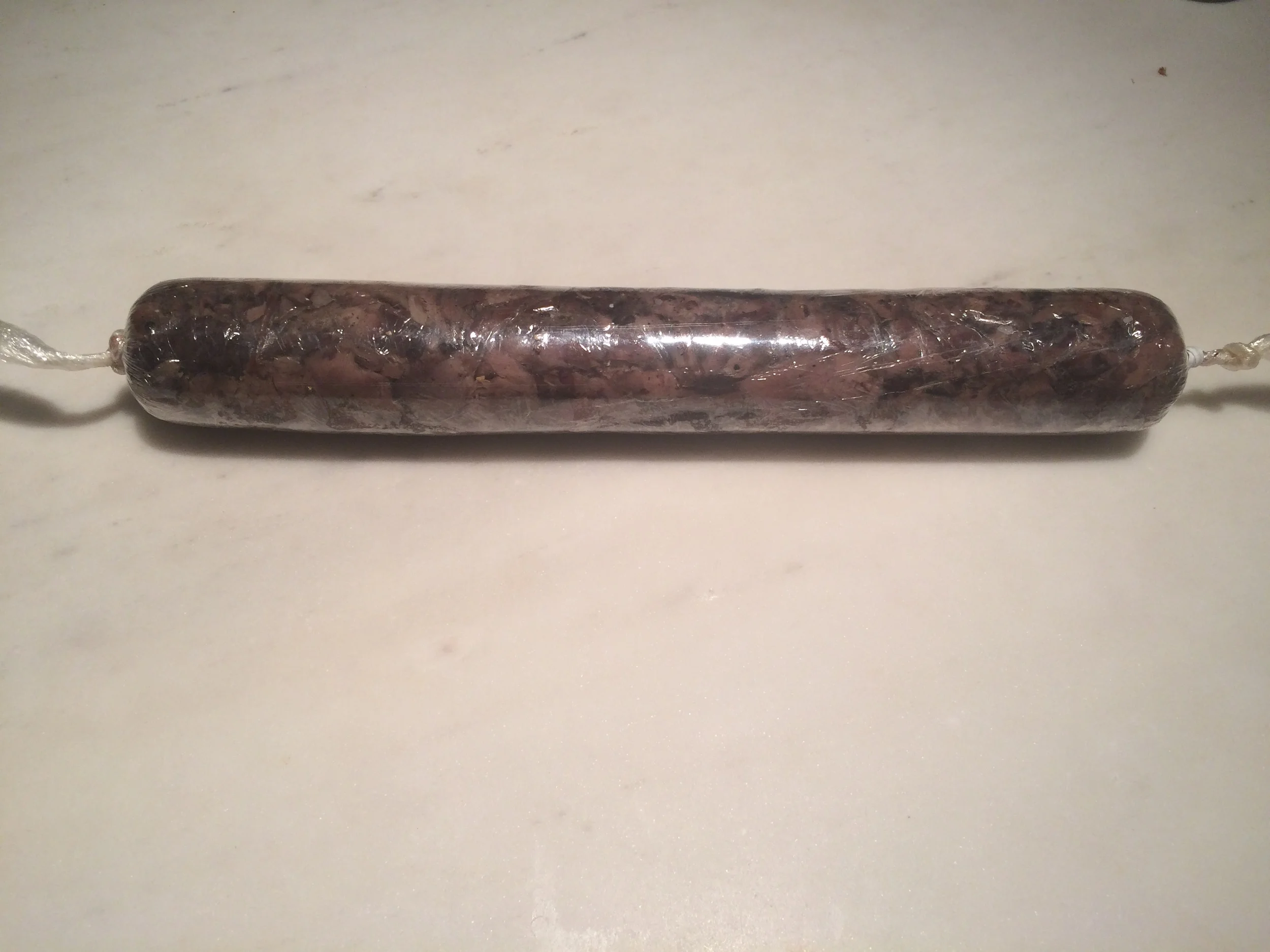 Quando acabar de enrolar, segure nas laterais de plástico que sobraram e arraste o rolo sobre a superfície, trazendo-o em sua direção. Ele vai enrolar estas extremidades e apertar ainda mais o a carne desfiada que está dentro. Isso é importante para que todos os espaços de ar entre as lascas de carne sejam preenchidas e o medalhão fique íntegro depois de cortar. 