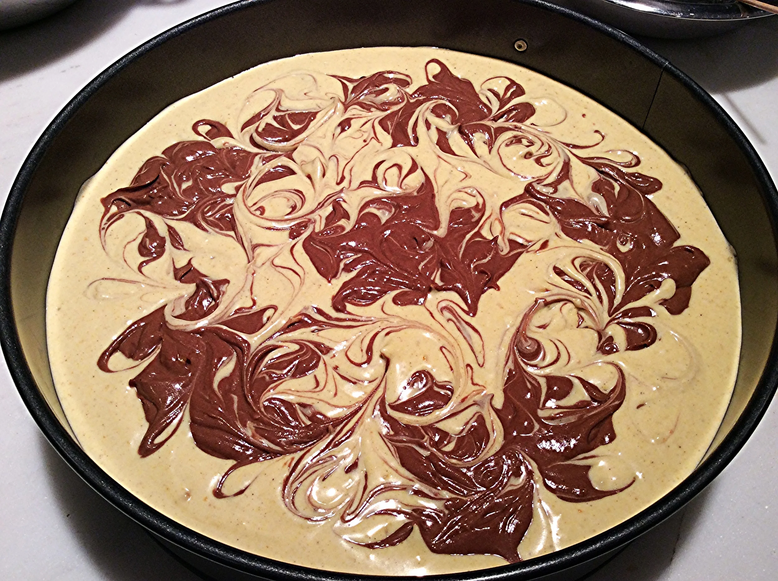 Aqui, a cheesecake pronta para ir ao forno da primeira vez que fiz. Na forma de cm que usei da segunda vez, a xícara reservada da massa de abóbora será adicionada às colheradas, como a de chocolate, nos espaços entre as porções marrons. Com isso, fazer o efeito marmorizado com o palito será bem mais interessante e próximo do desta foto. Para fazê-lo, divida a forma em 4 quadrantes imaginários. Afunde um palito para churrasco na massa do primeiro quadrante a ser trabalhado, com cuidado para não danificar a camada de biscoito. Com a ponta dele submersa, escreva a letra L cursiva várias vezes, para que as duas cores de massa se entrelacem, criando desenhos. Quando achar que já está bom neste quadrante, gire a forma e repita o processo. Assim sucessivamente, até que termine. Dê algumas leves batidas da forma contra a bancada, pra assentar a massa e eliminar possíveis bolhas de ar. 