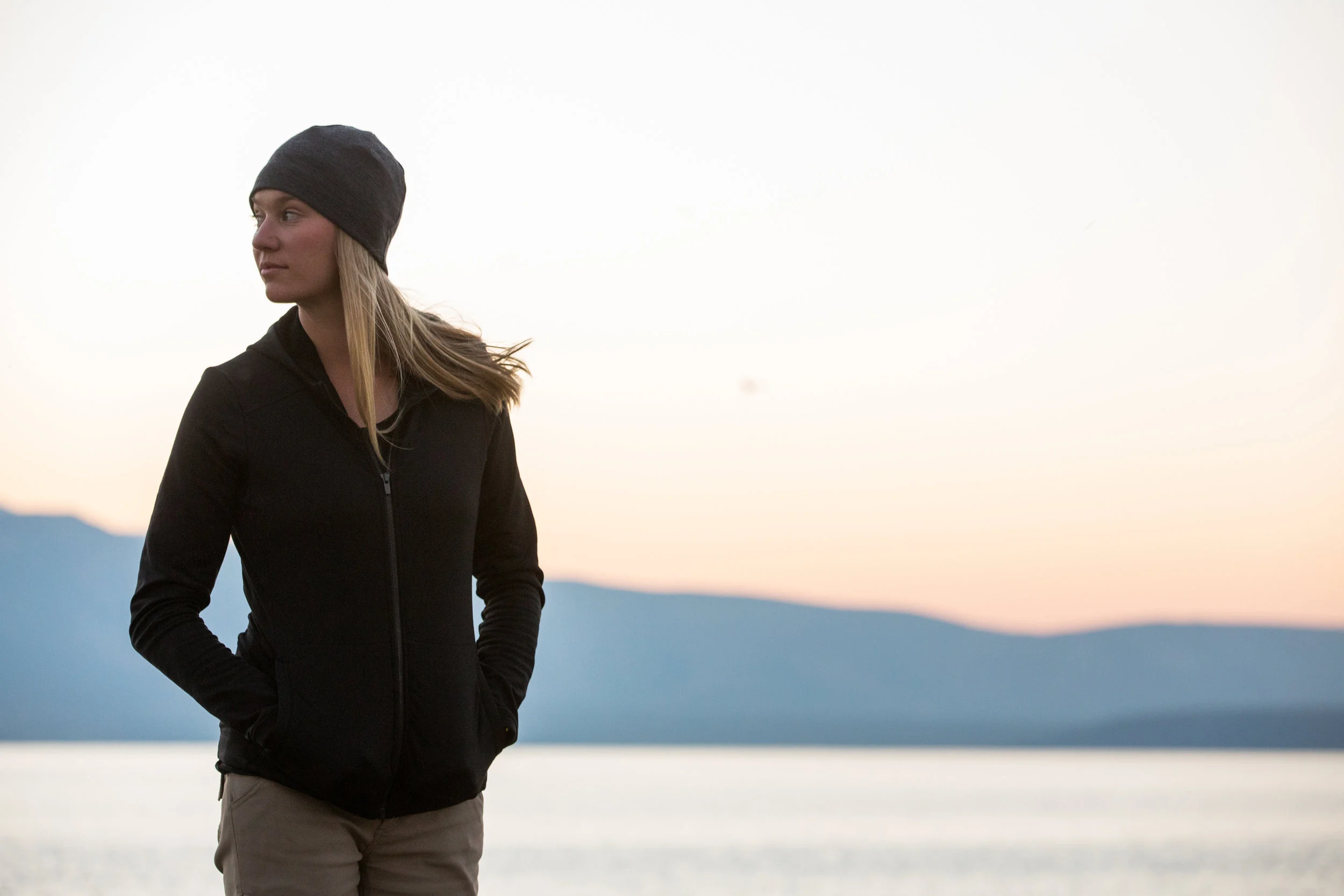  Young woman stands in front of mountain and lake landscape at sunset. 