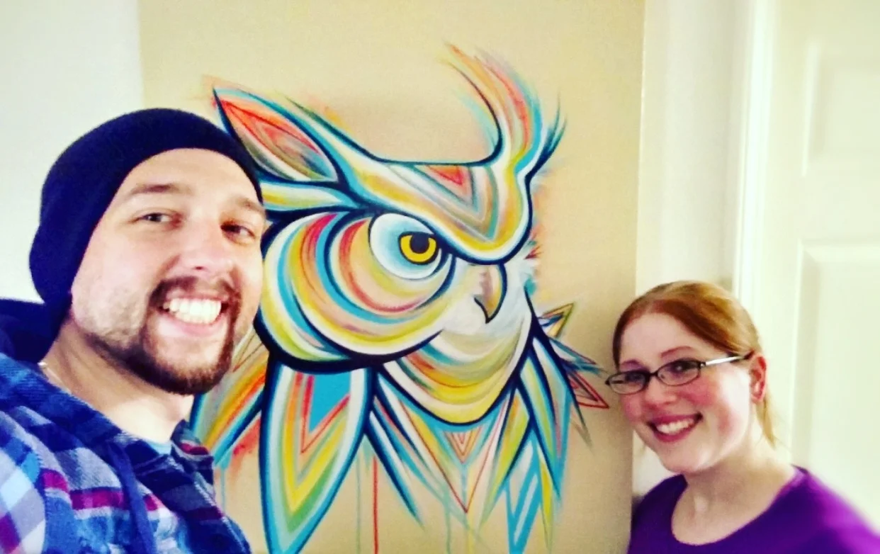 Image of a smiling couple with a large colorful geometric owl painting