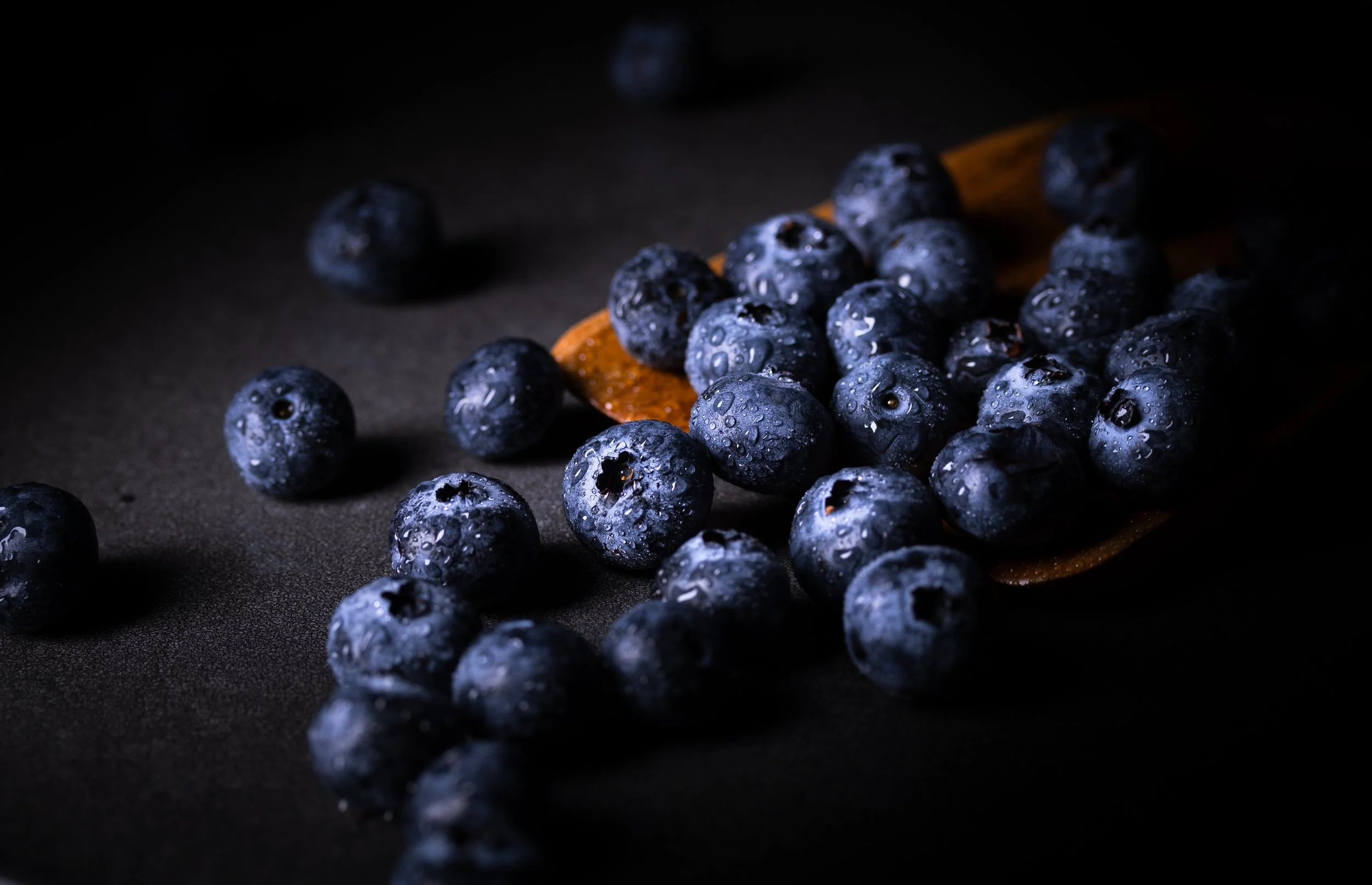 Close-up of blueberries with water droplets on a dark surface, some on a wooden spoon.
