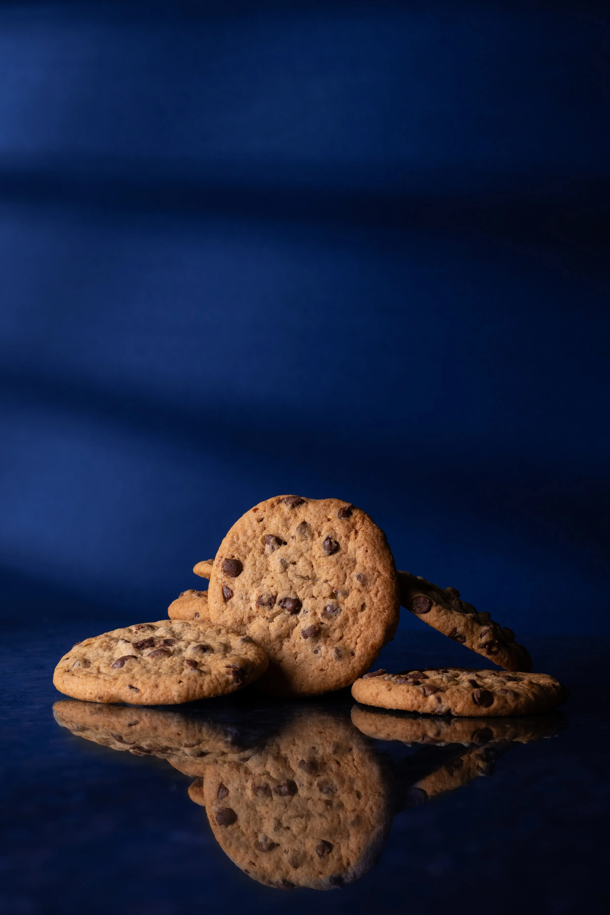 Close-up of chocolate chip cookies stacked against a dark blue background, with their reflection shown on a shiny surface.