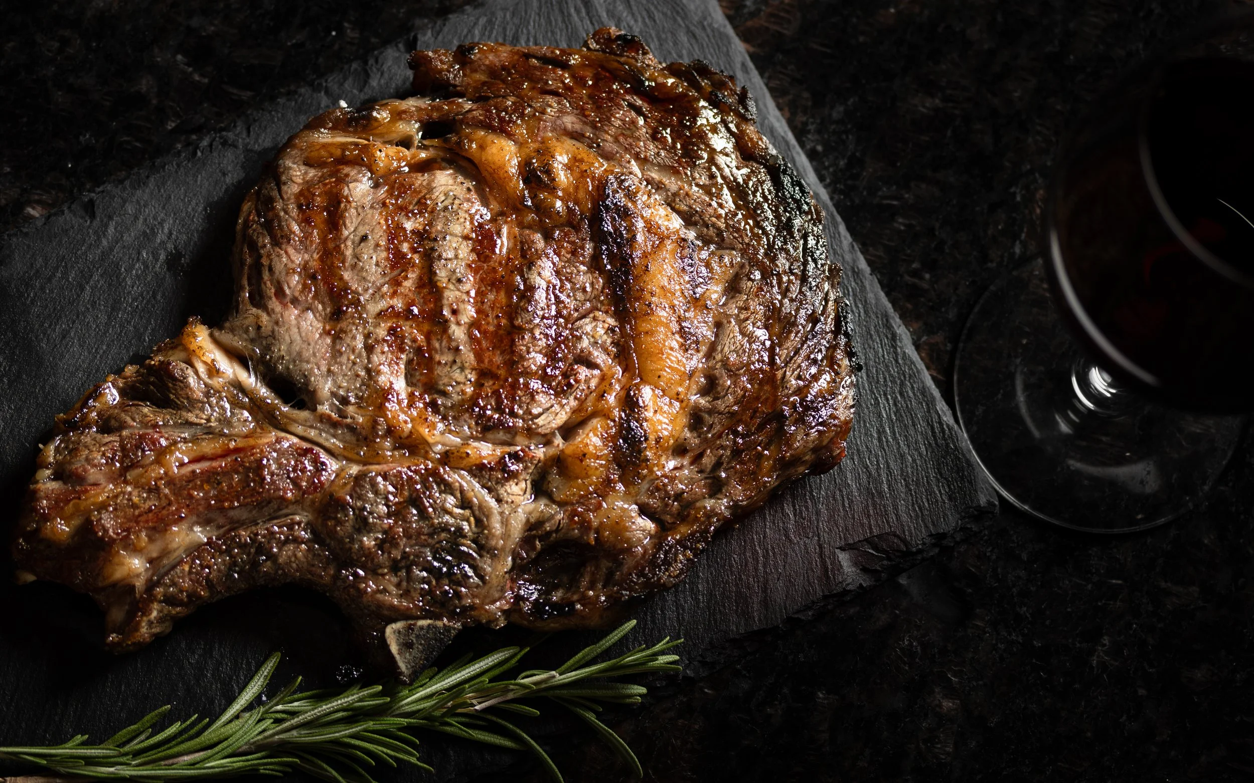 A cooked steak on a black slate serving board with rosemary sprigs beside it.