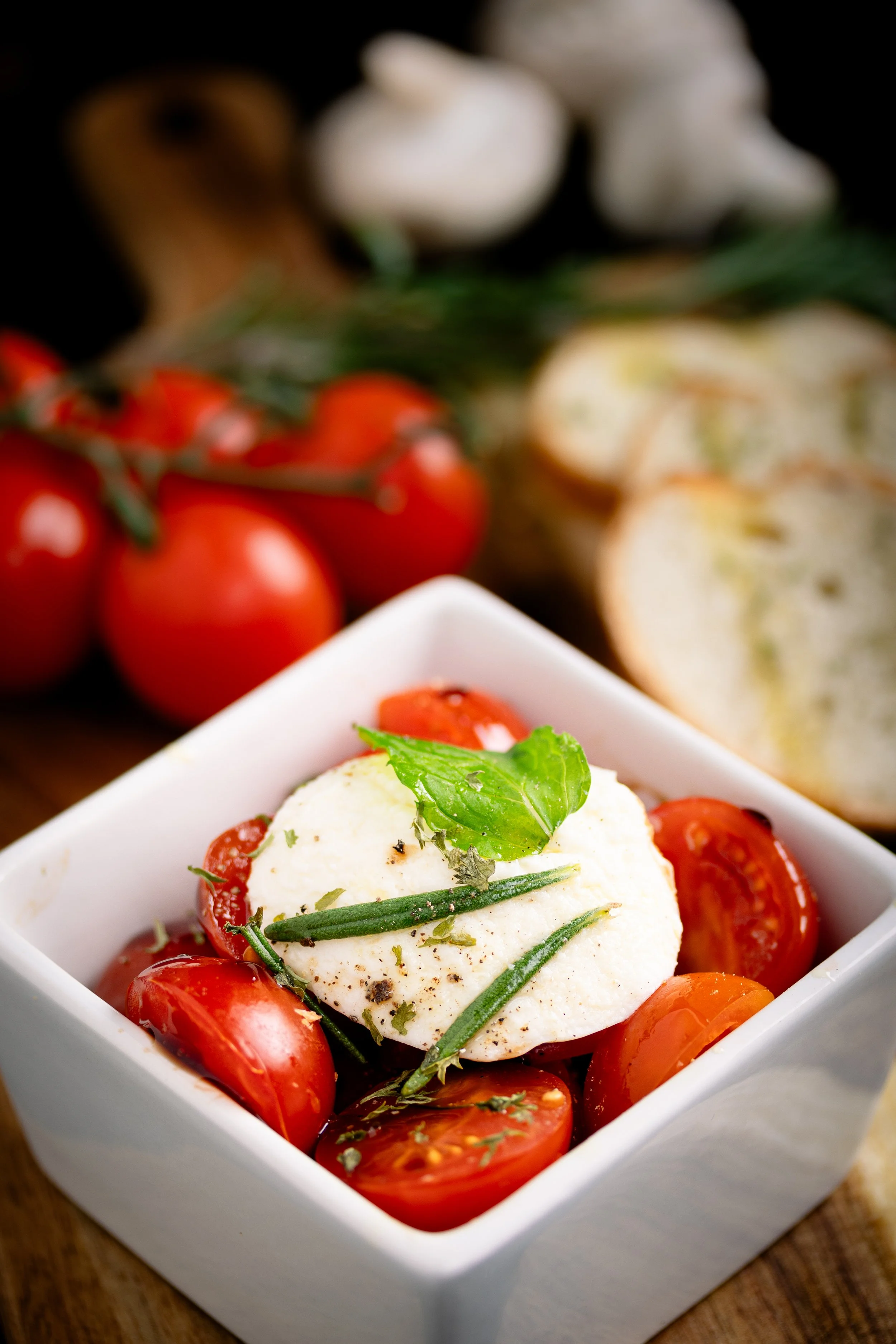 A small square white bowl filled with cherry tomatoes, topped with a ball of fresh mozzarella cheese garnished with basil leaves and rosemary sprigs. In the background, there are blurrily visible garlic and onions.