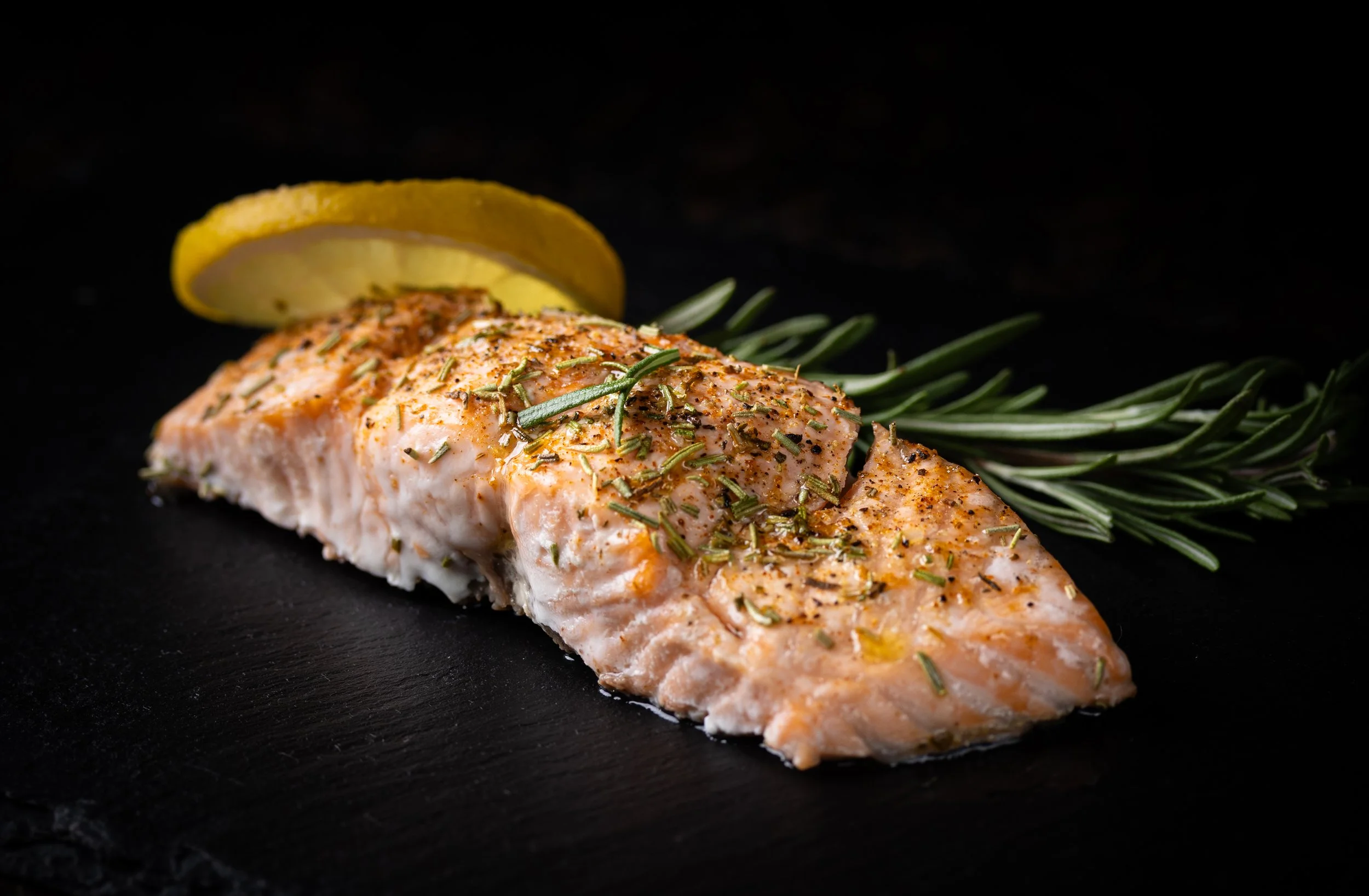Cooked salmon fillet seasoned with herbs, garnished with rosemary sprigs, with a lemon wedge on a dark surface.