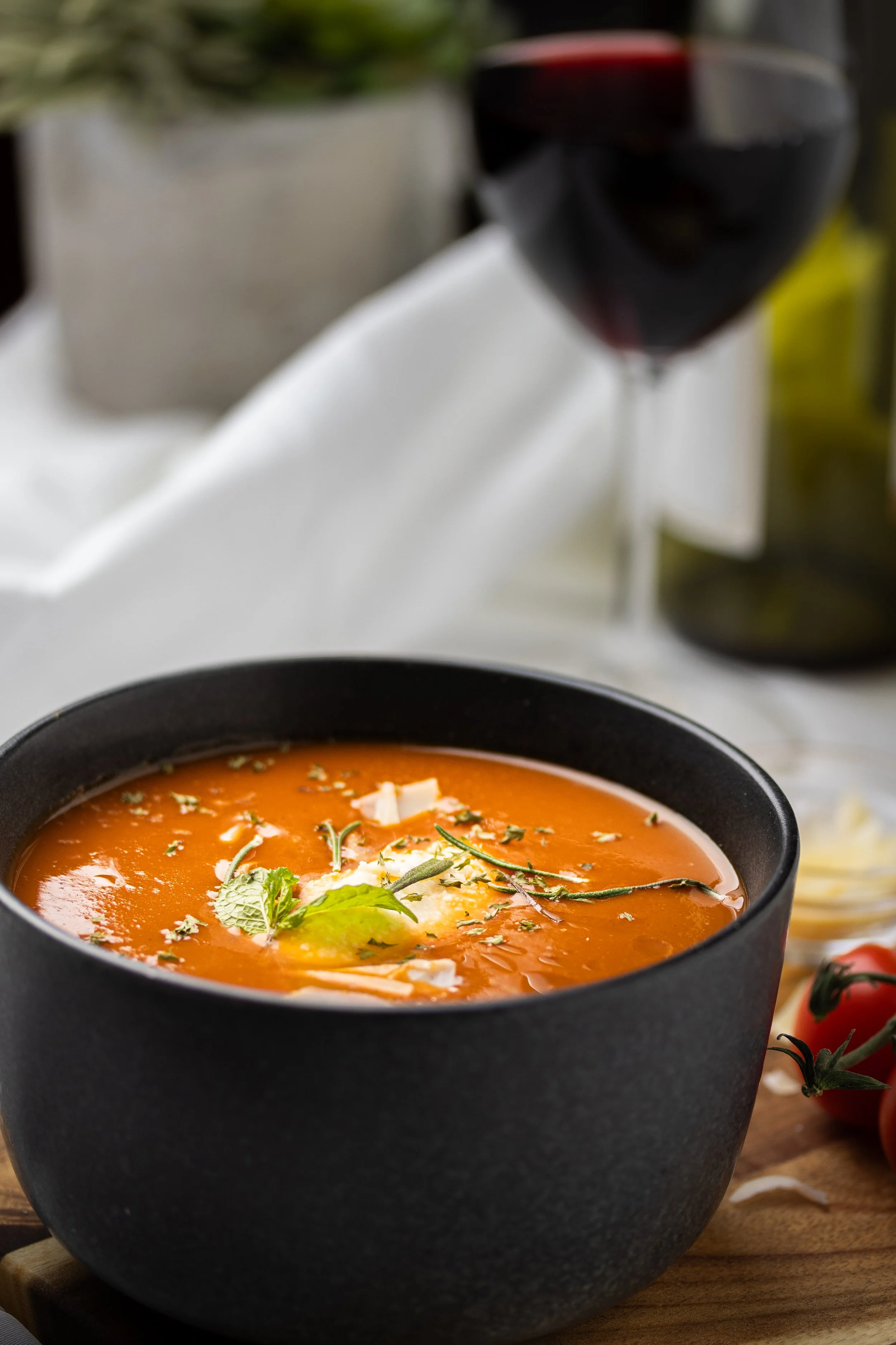 A bowl of tomato soup garnished with herbs and a slice of cheese, with a glass of red wine in the background.