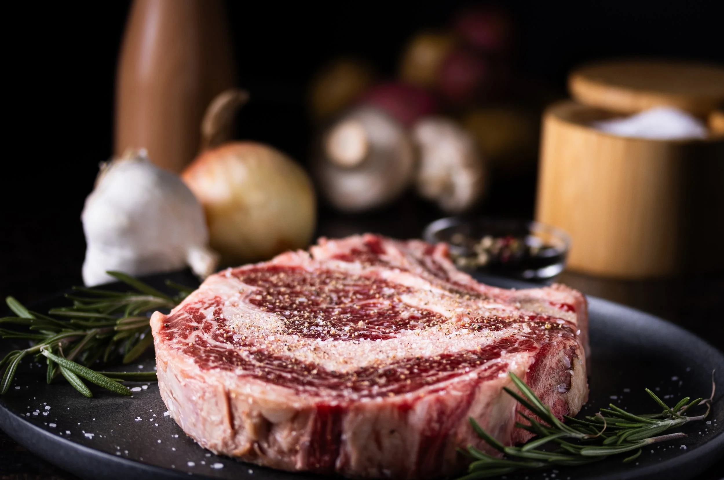 A raw marbled steak seasoned with salt and pepper, garnished with sprigs of rosemary, on a dark surface with garlic and a wooden container in the background.