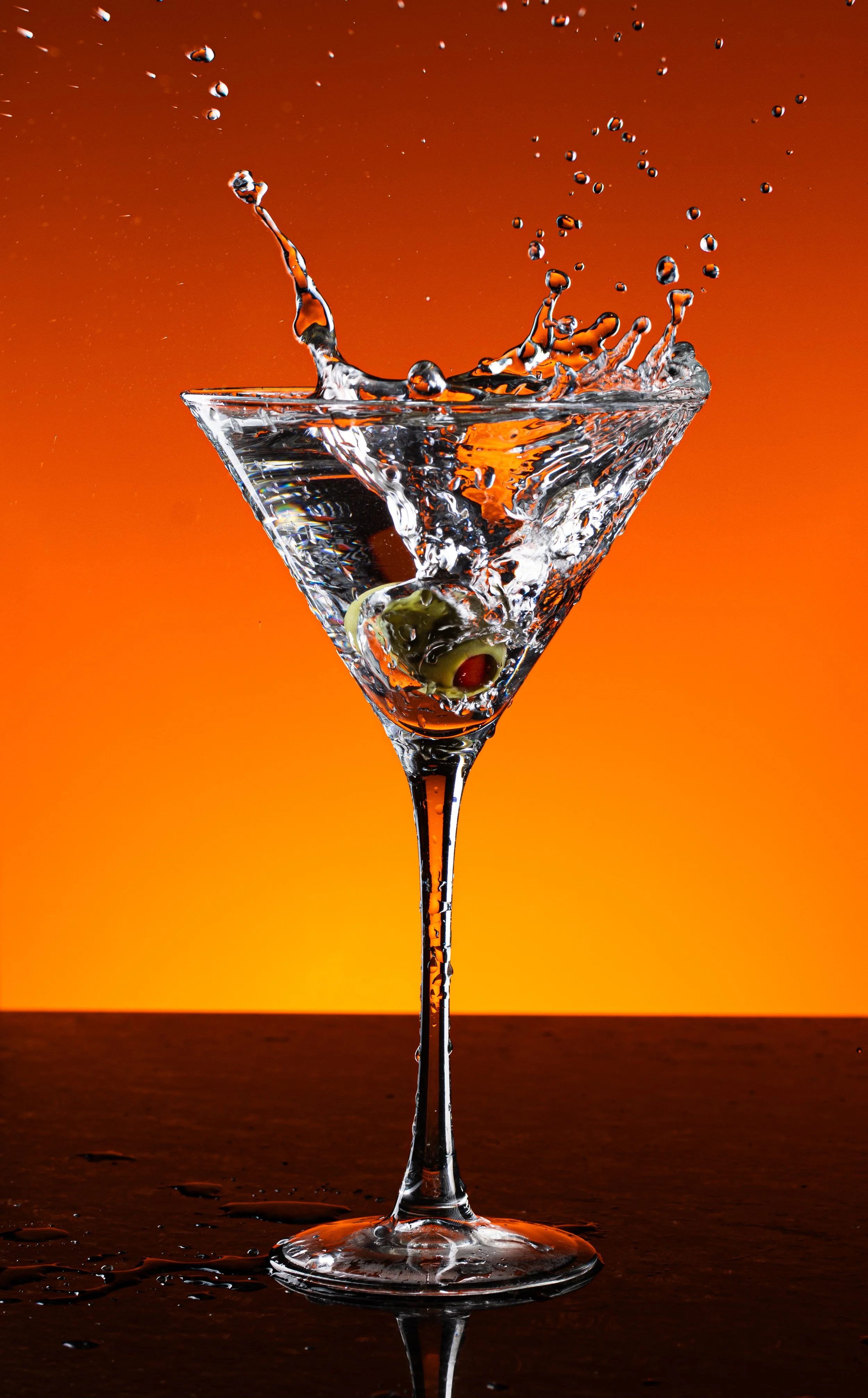 A martini glass with olives inside, filled with water, with water splashing out as it is disturbed, against an orange gradient background.