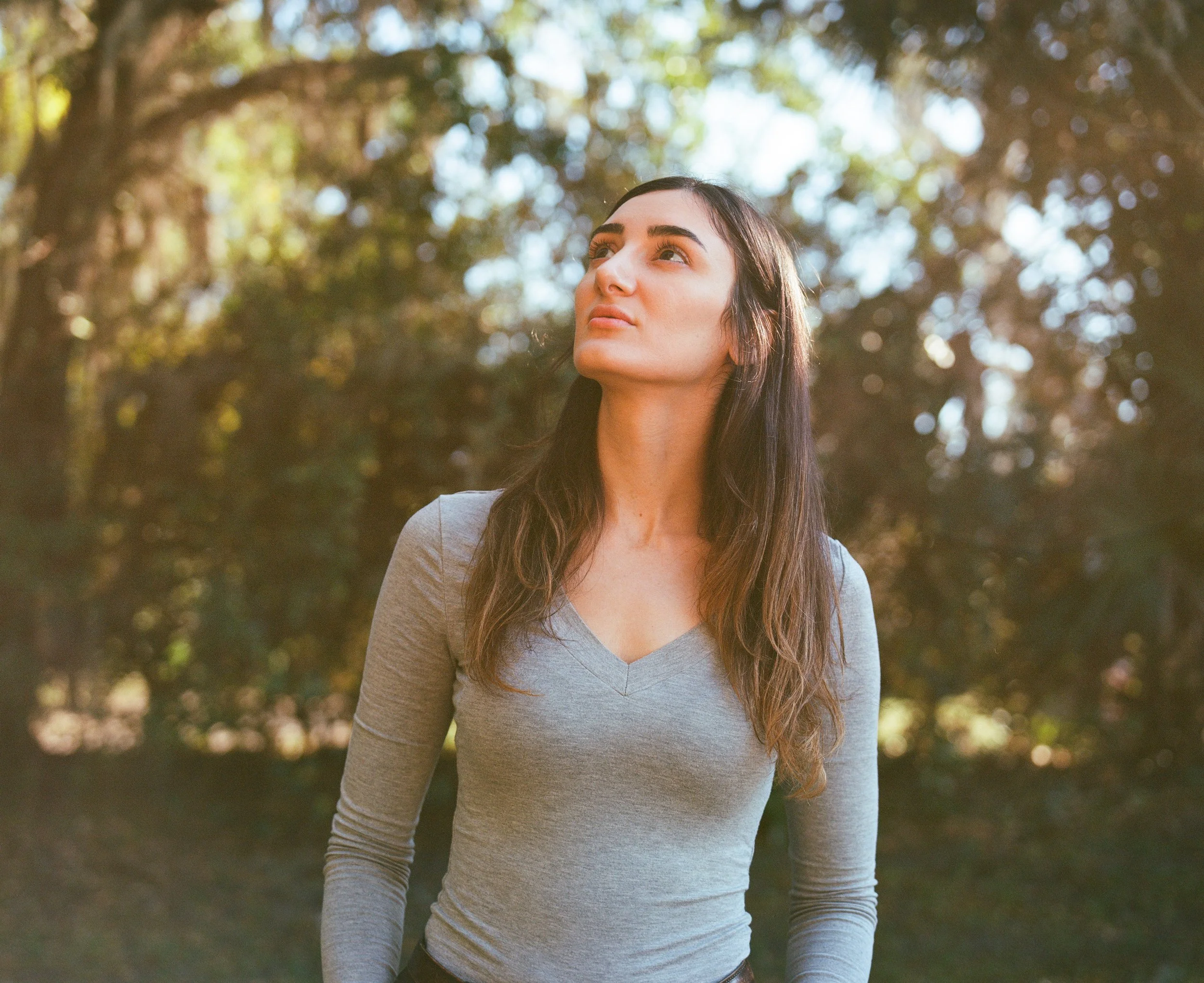 A medium shot portrait of tattoo artist Nicki Giustine standing outdoors, looking upward with a thoughtful expression against a background of soft, blurred green trees and sunlight.