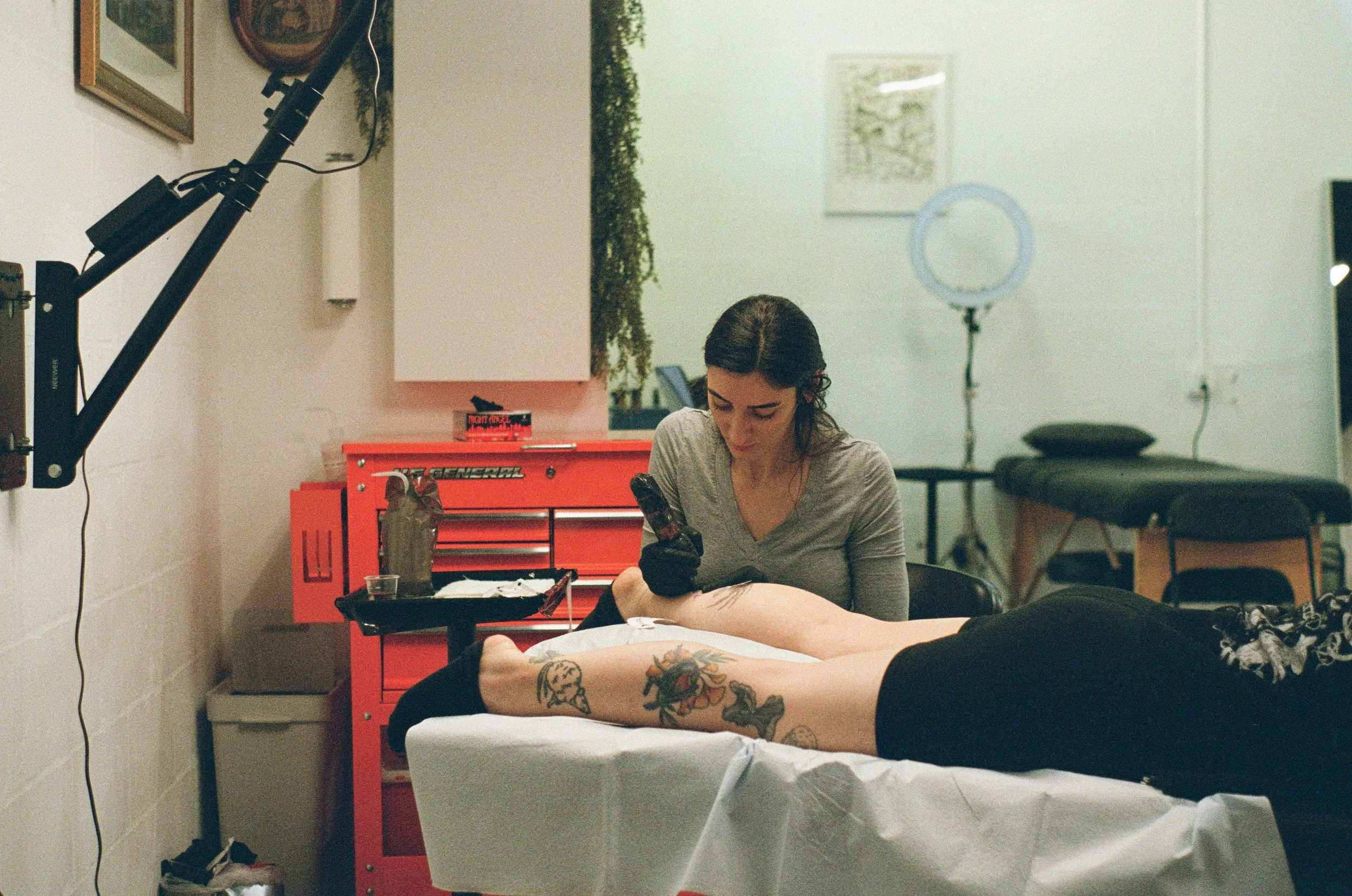 A tattoo artist works at her station inside a well-lit creative studio, leaning over a client on a table.