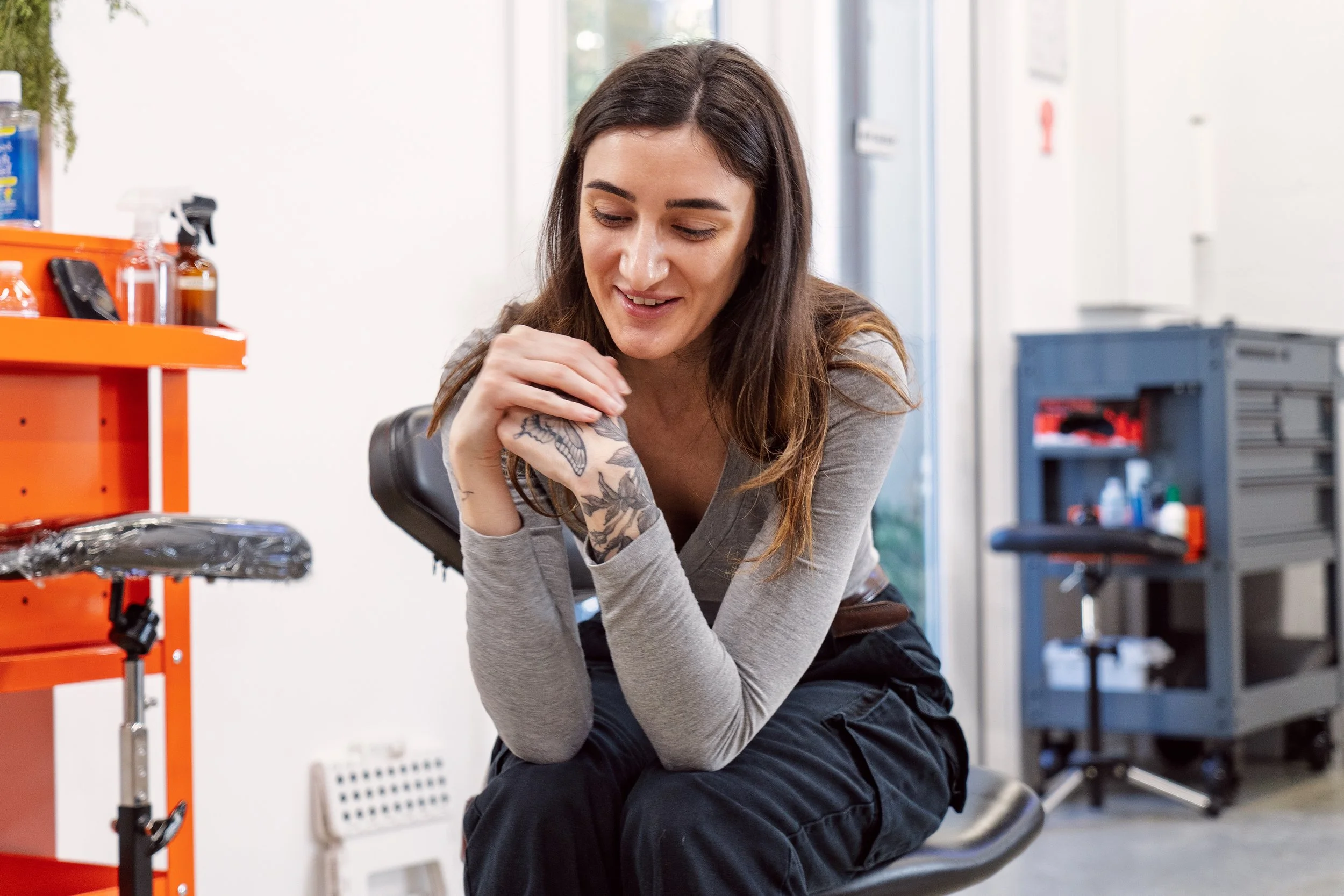 A close-up of tattoo artist Nicki Giustine sitting on a stool and smiling warmly while looking at her client's arm during a break in the session.