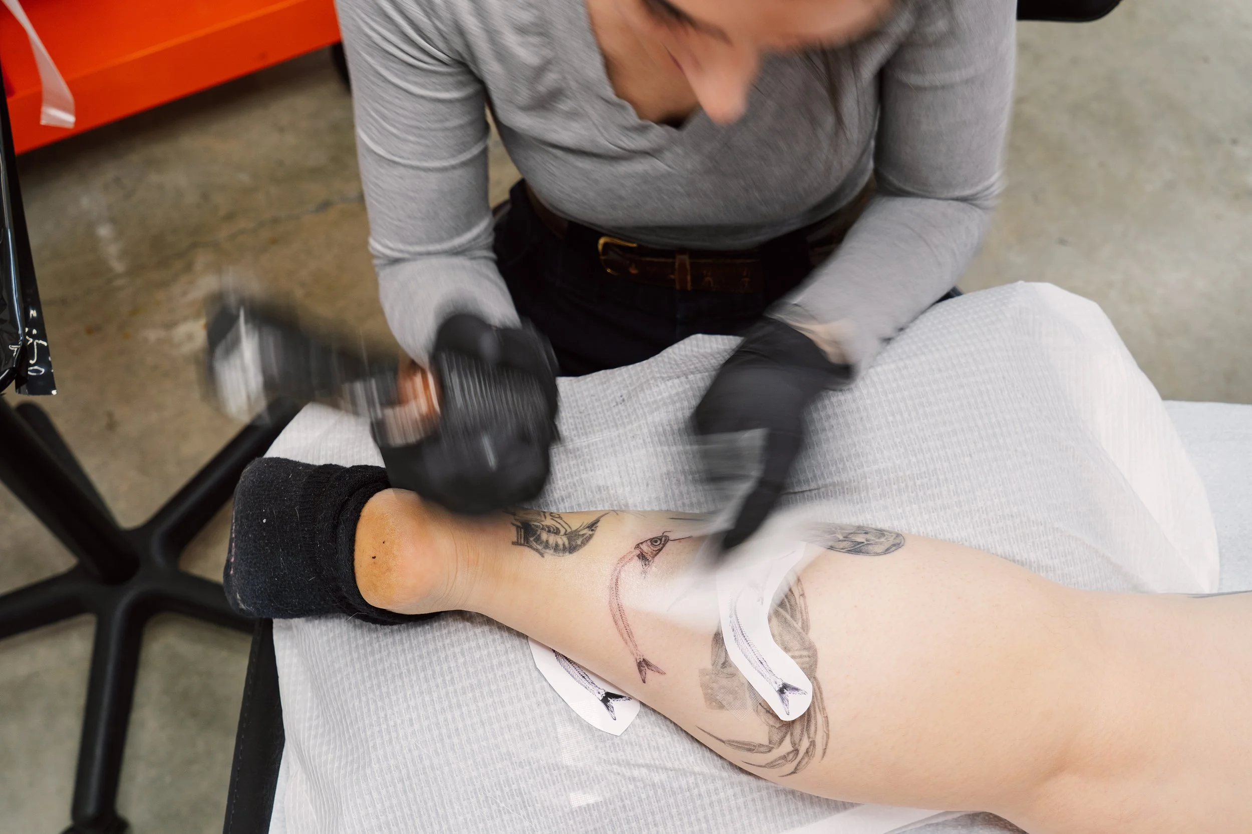 A close-up of tattoo artist Nicki Giustine wearing black gloves as she uses a tattoo machine to carefully apply detail to a client's arm.