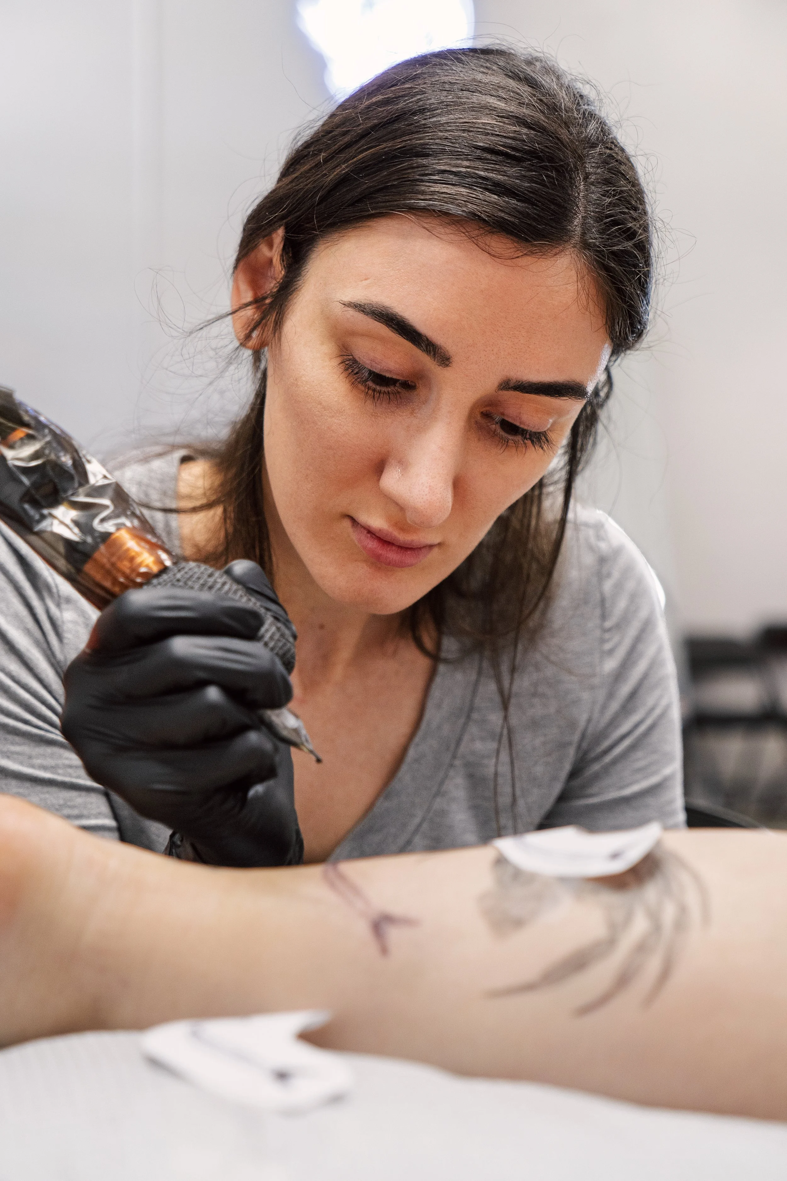 A woman with brown hair leans over a leg with a tattoo machine.