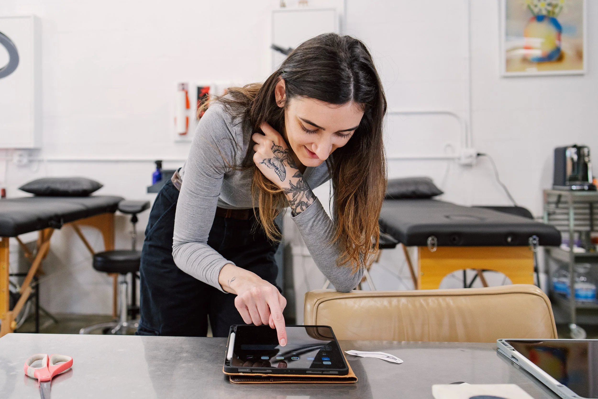 A woman leans over a table while pointing at an iPad. Tattoo beds and artwork fill the background.