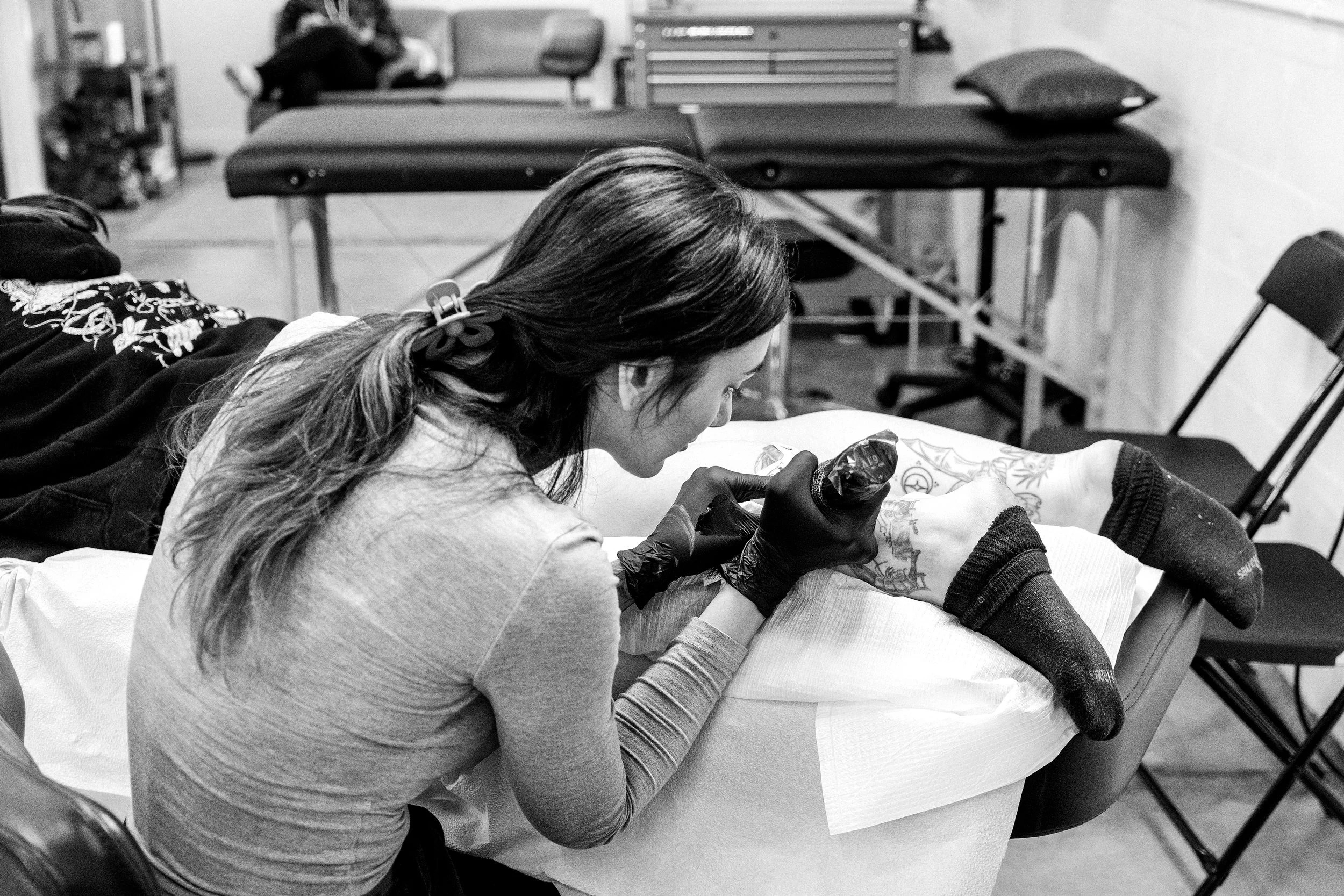 A woman holding a tattoo machine leans over a client's leg on a table.