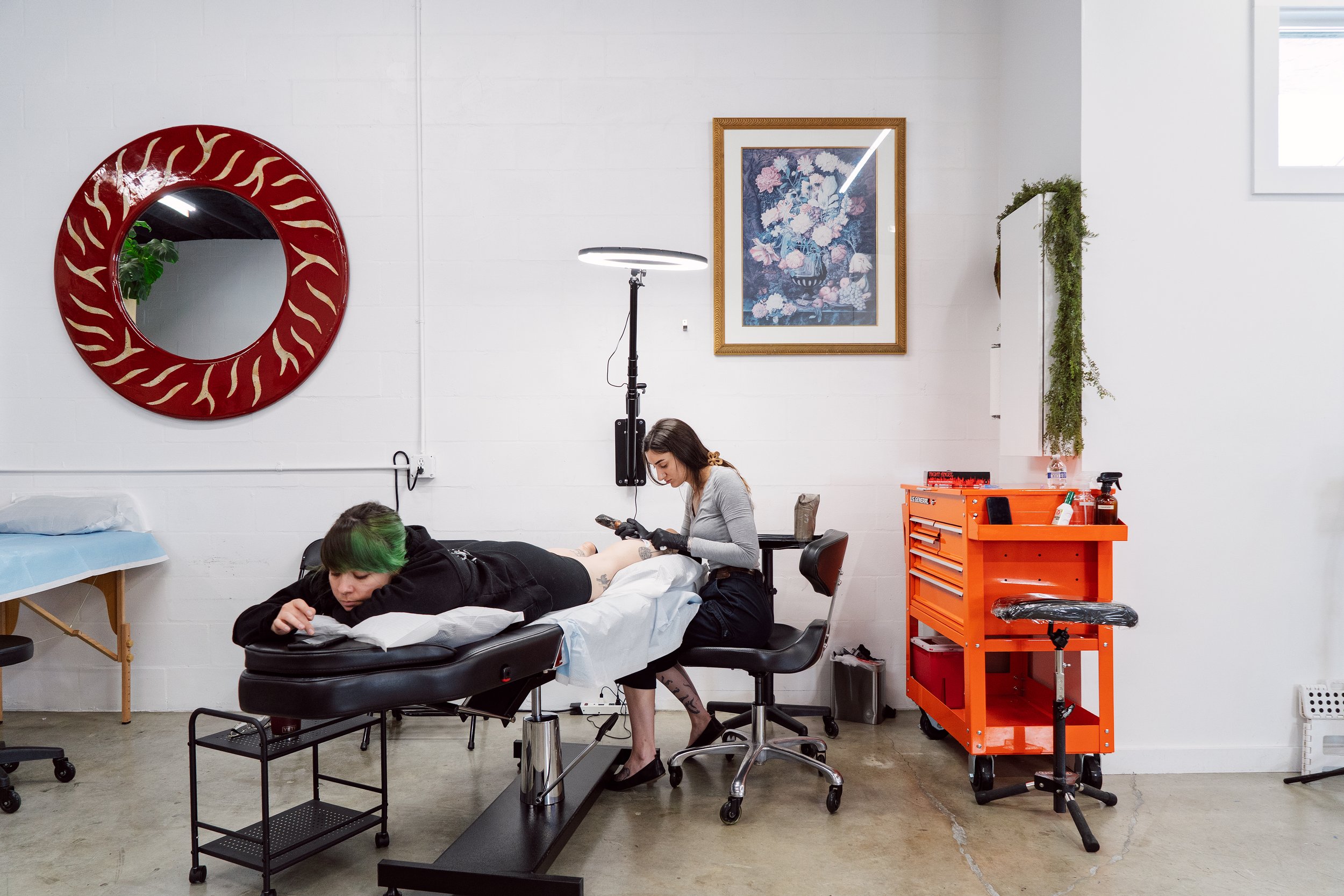 Tattoo artist at work in a bright studio with an orange tool chest and a large round mirror on the wall.