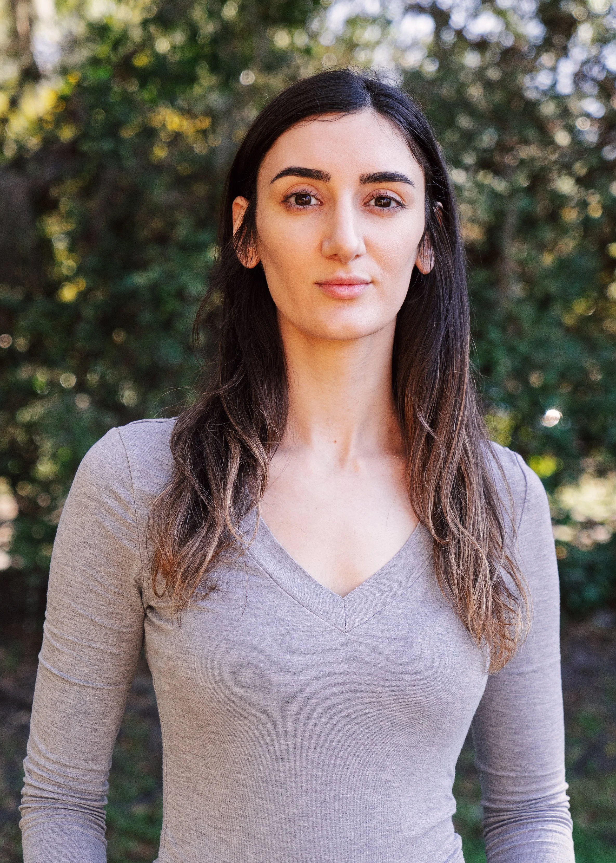 A woman with long brown hair and a gray sweater stands in front of a blurred green background.