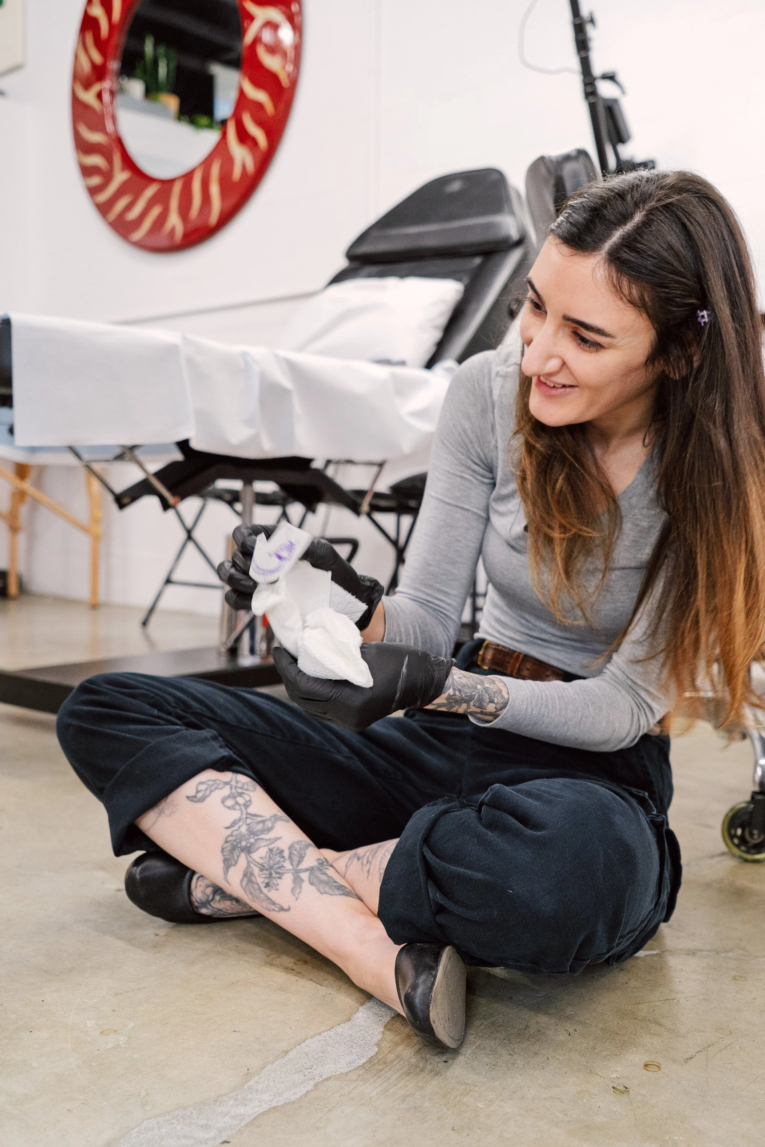 A smiling woman sits cross legged on a tattoo studio floor
