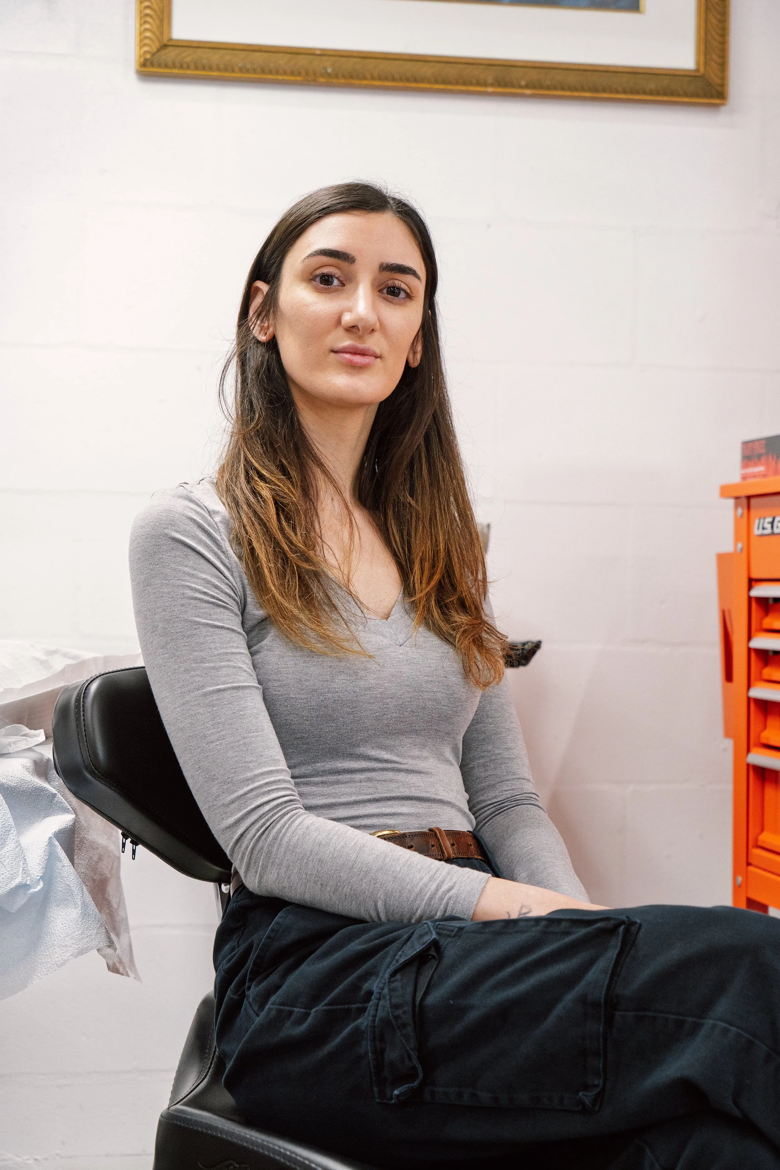 A portrait of tattoo artist Nicki Giustine sitting on a black studio chair, looking directly at the camera with a calm expression.