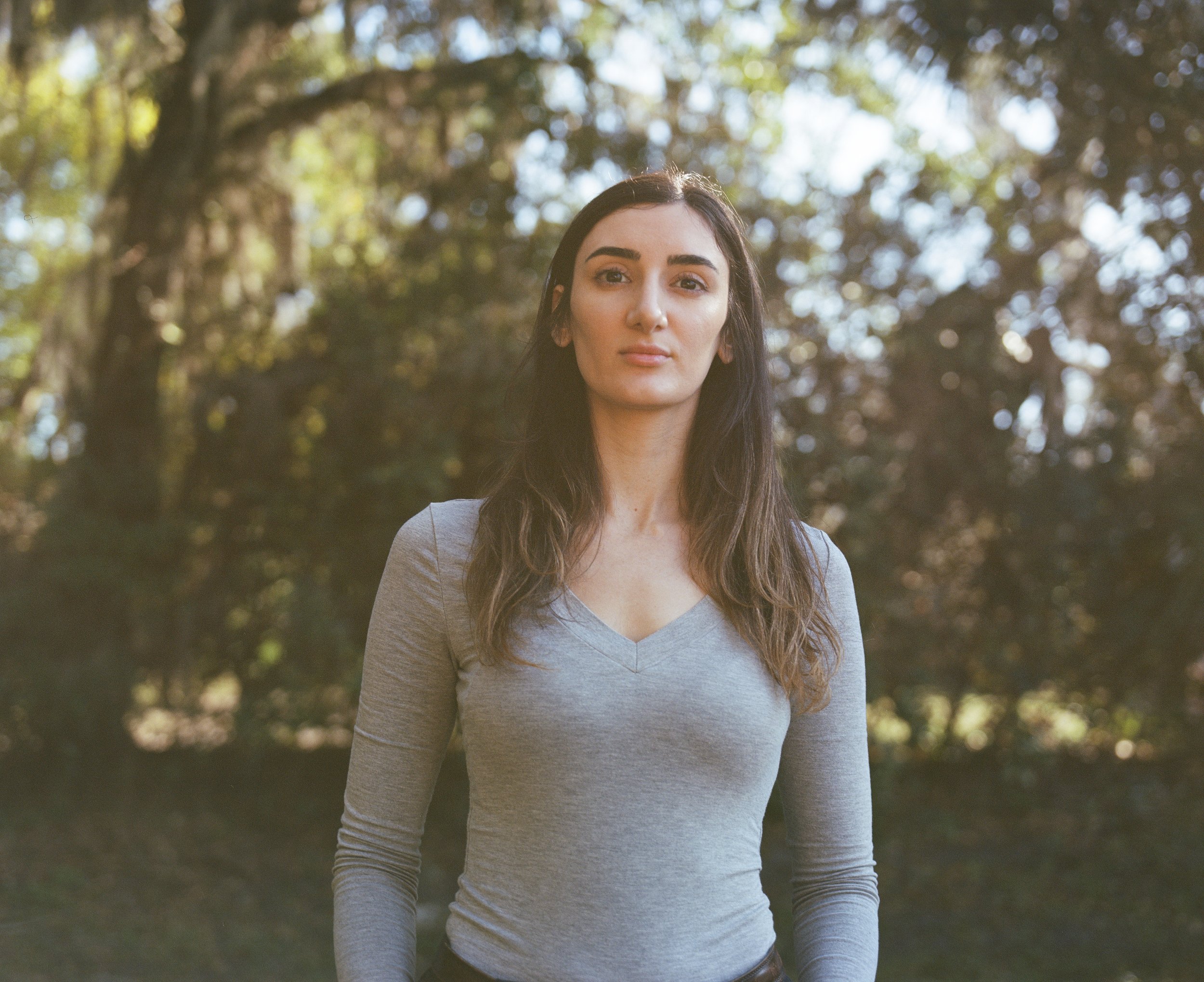A woman with long hair in a gray long sleeve shirt stands in front of a blurred forest.