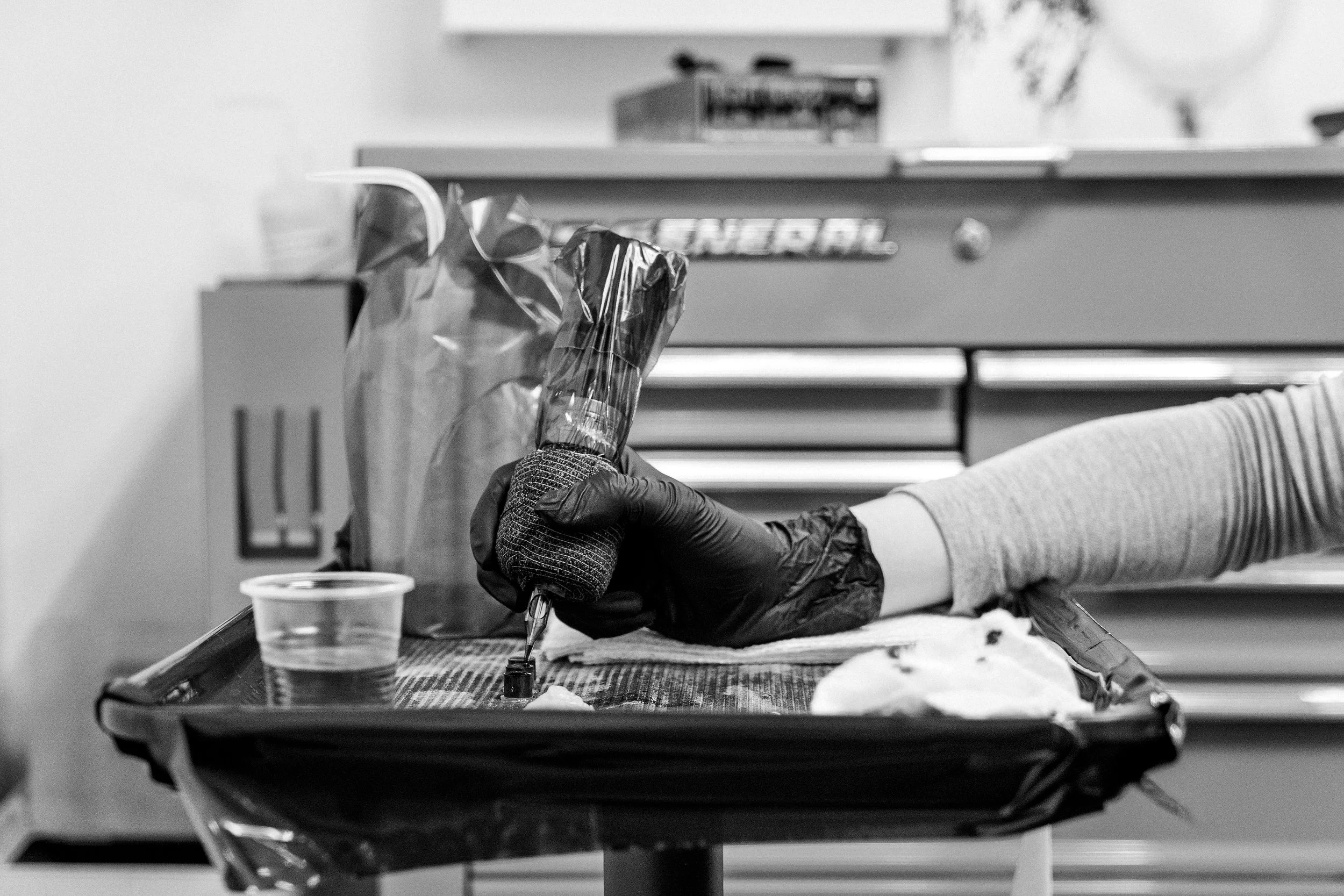 A hand holding a tattoo machine over a small pot of ink on a tray filled with other items.