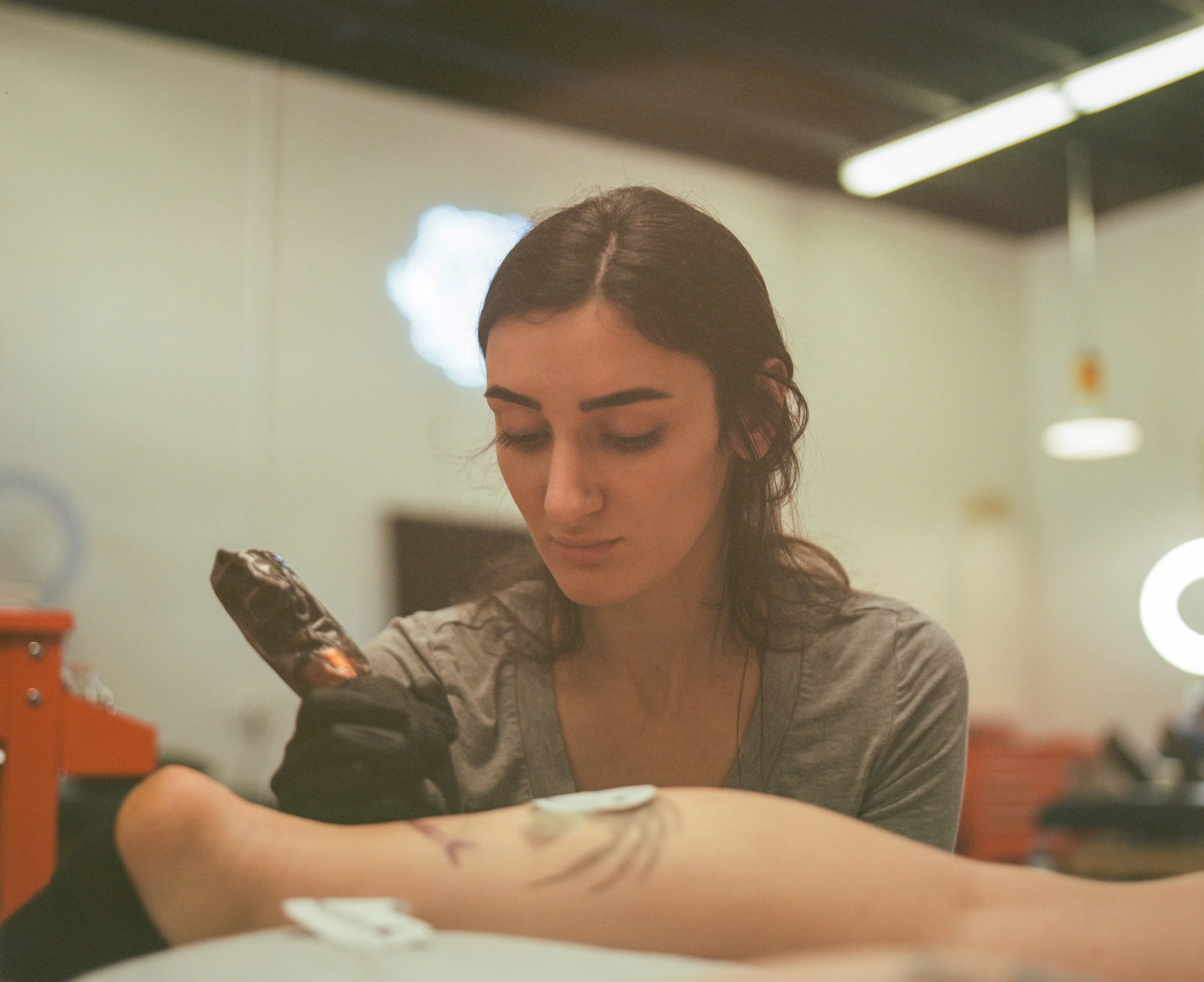 A tattoo artist with long dark hair concentrates on her work as she tattoos a client's leg in a brightly lit studio.