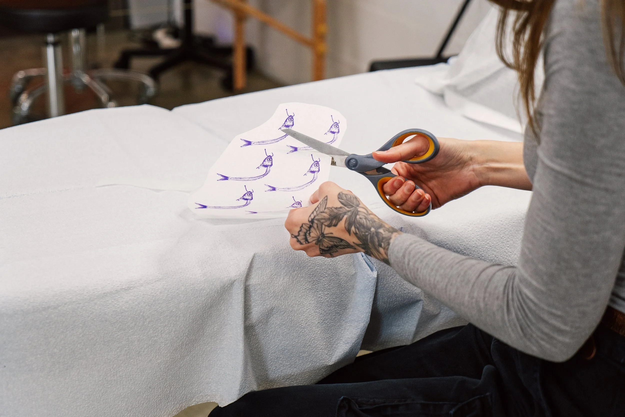 A close-up of a tattoo artist's tattooed hands using scissors to precisely cut out printed custom tattoo stencils on a white table.