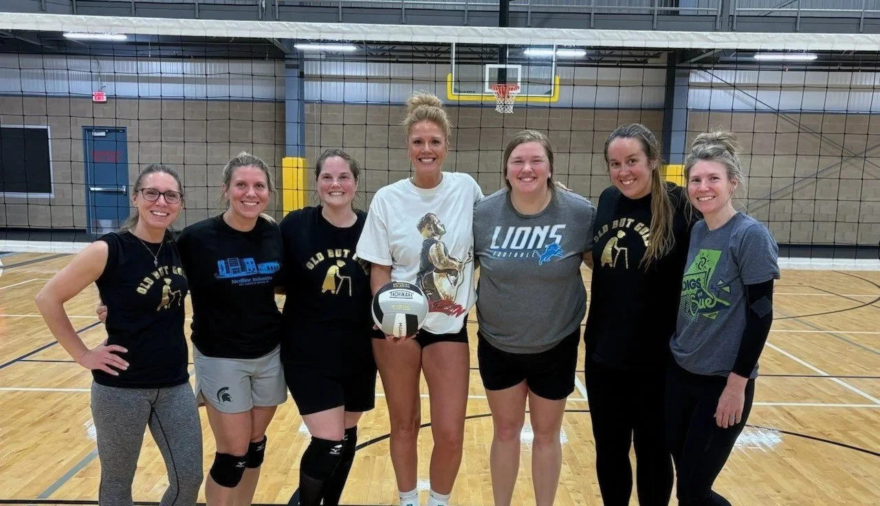 A group of women pose in front of a volleyball net with a volleyball.