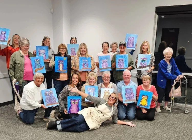 A group of participants pose with their paintings of colorful owls.