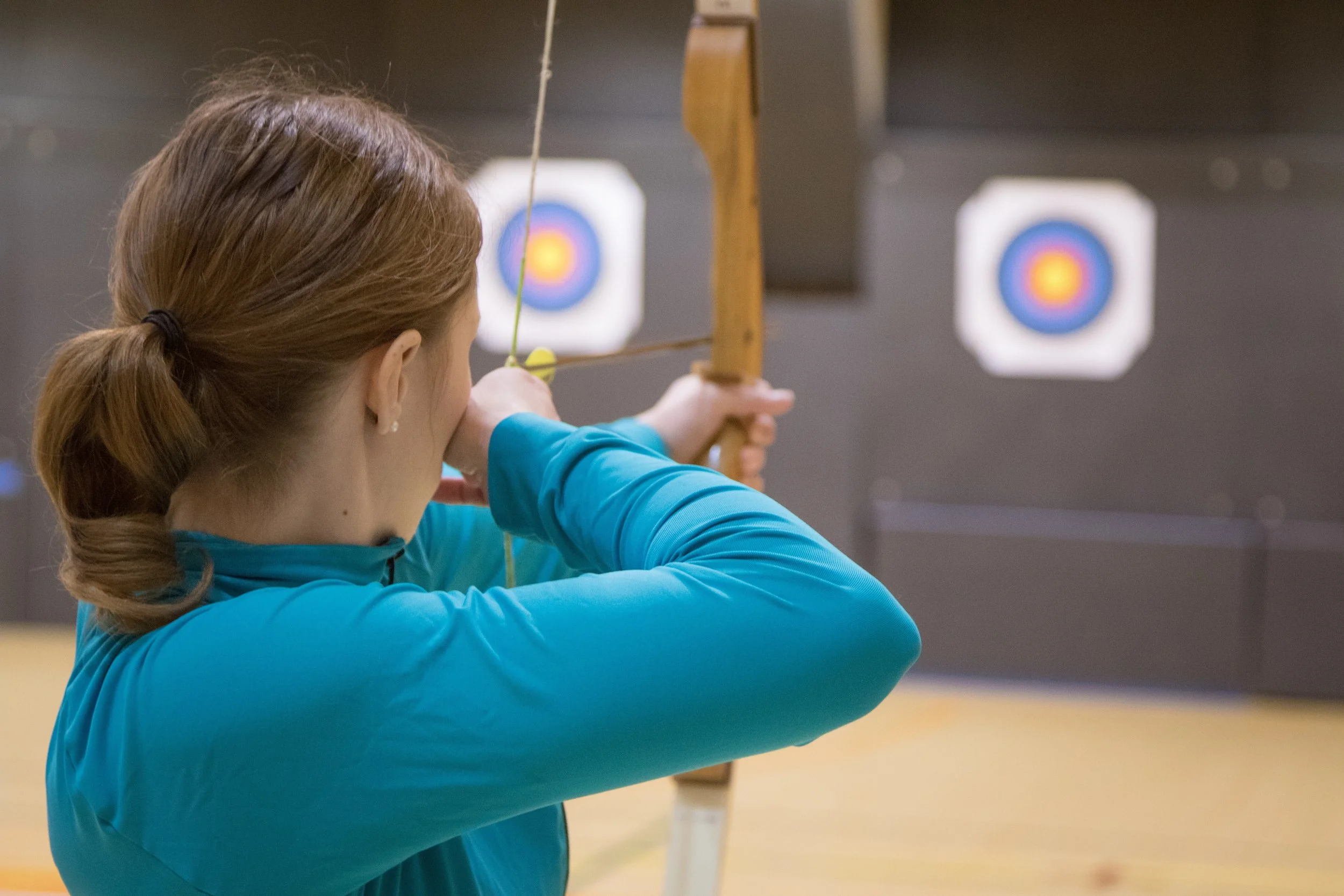 A young woman aims a bow and arrow at a multicolored target at an indoor range.