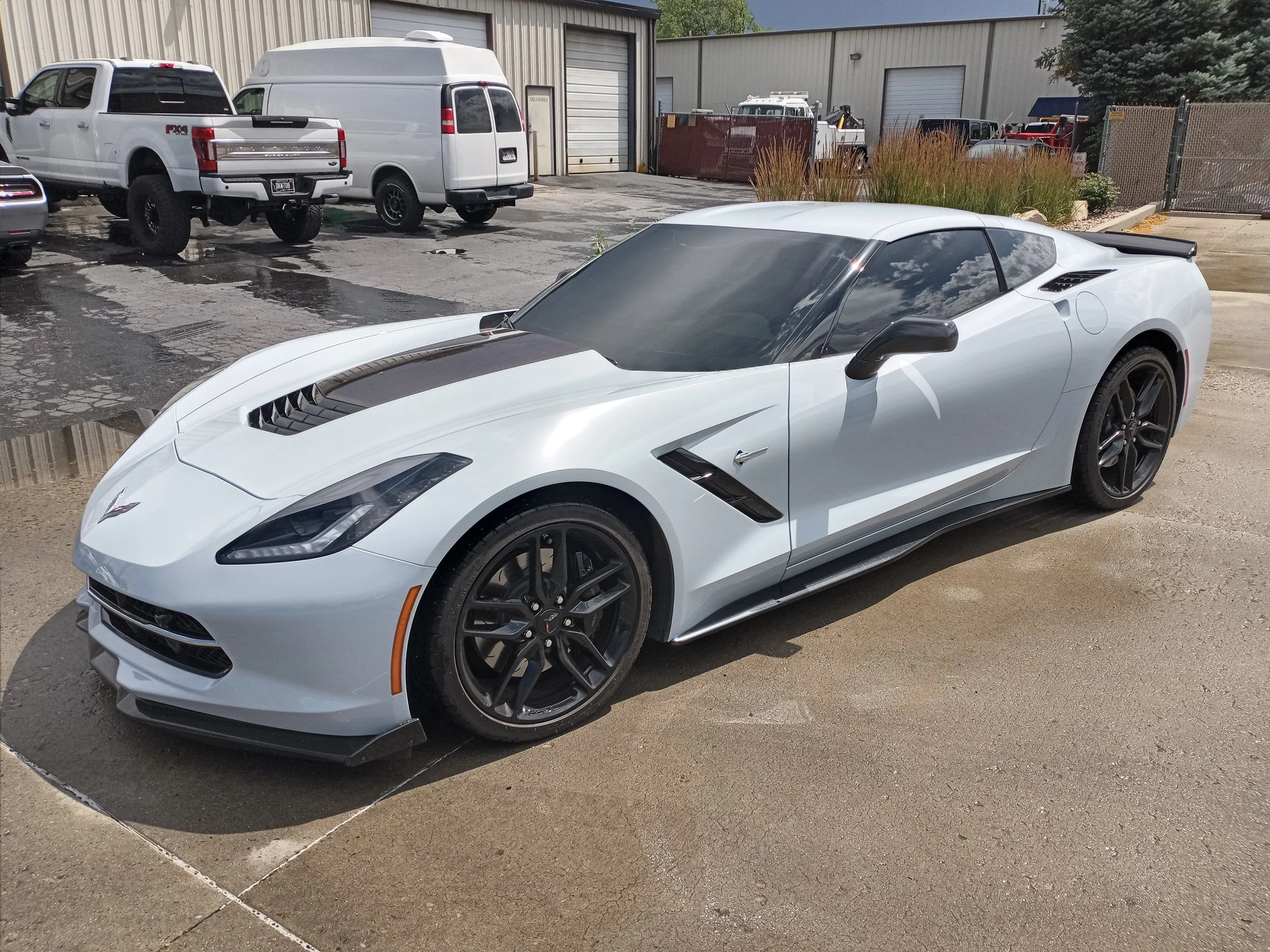 A white sports car parked in an outdoor lot, next to other vehicles including a black and white pickup truck and a white van, with industrial buildings in the background.