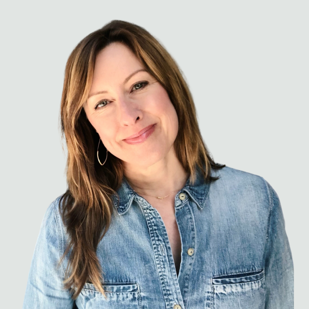 A woman with shoulder-length brown hair, wearing a denim shirt and hoop earrings, smiling at the camera against a plain light background.