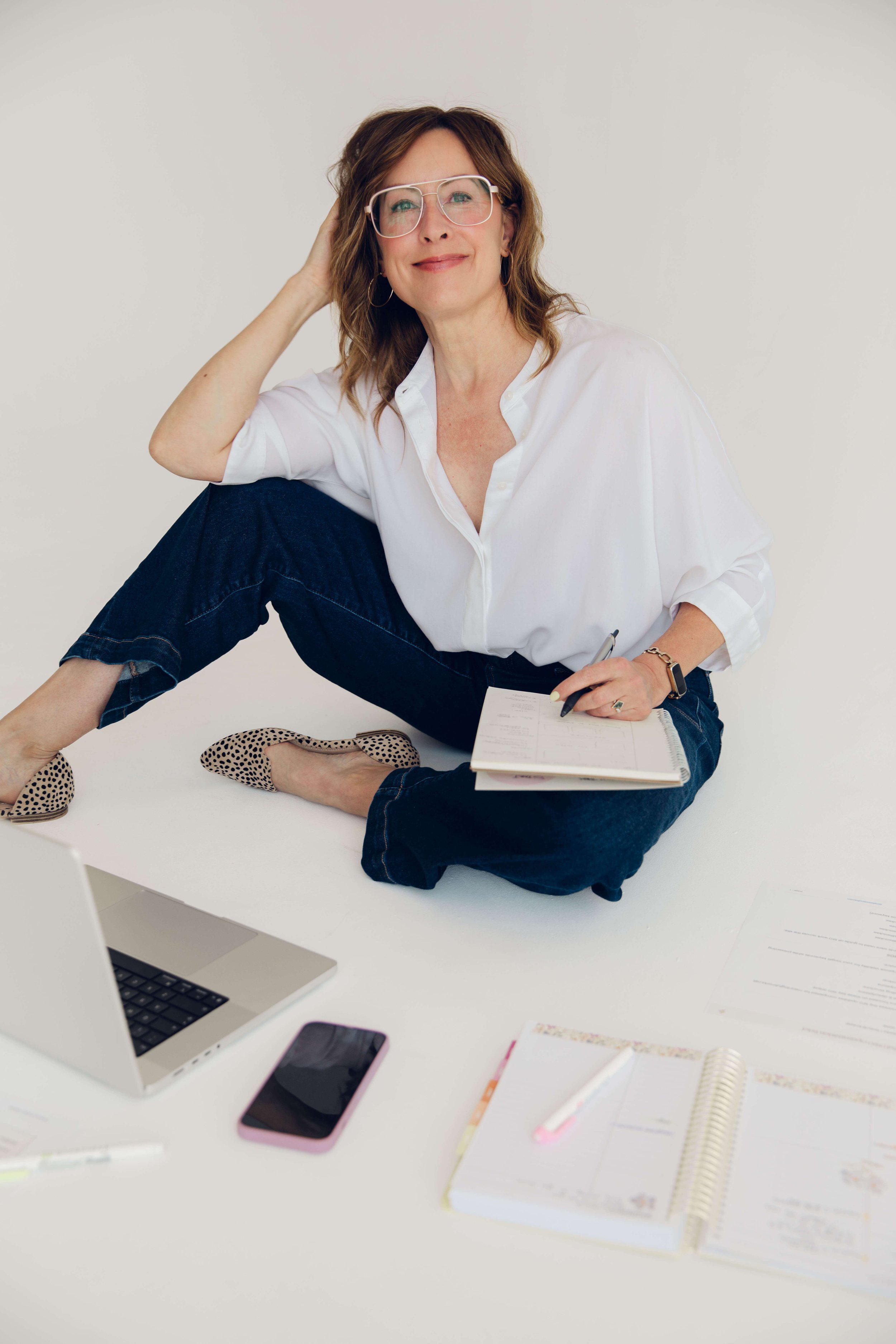 Website designer for therapists, Deana Ward, is holding a pen and notebook. Items on the desk include a laptop, smartphone, notebooks, and pens.